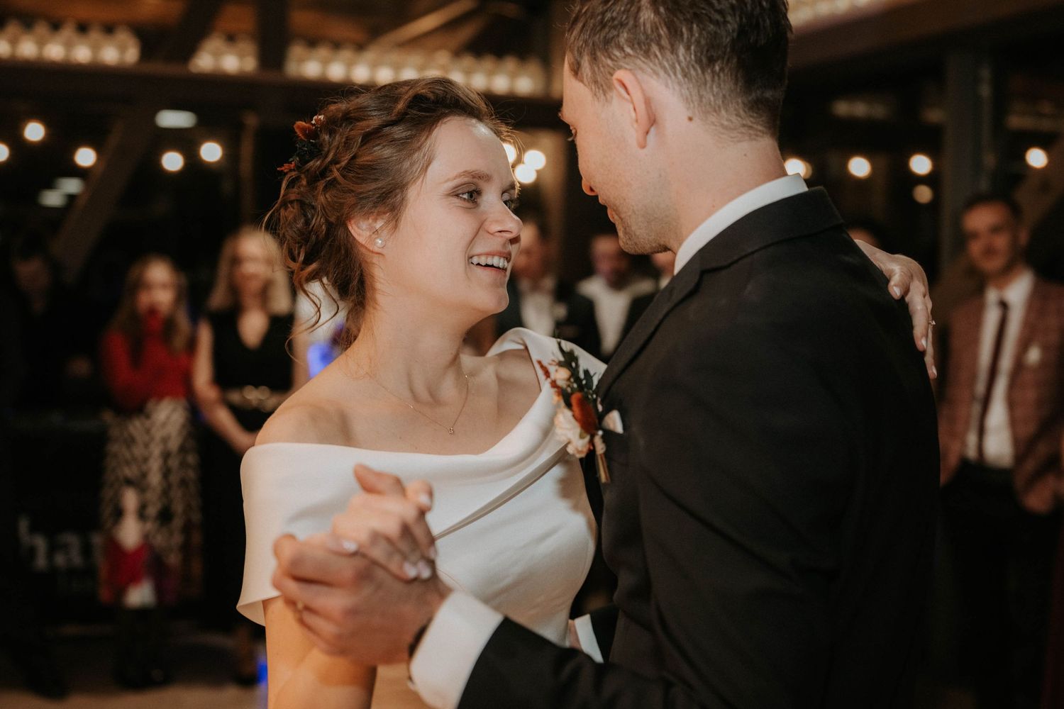Bride and groom share their first dance, smiling joyfully at wedding reception with guests in background.