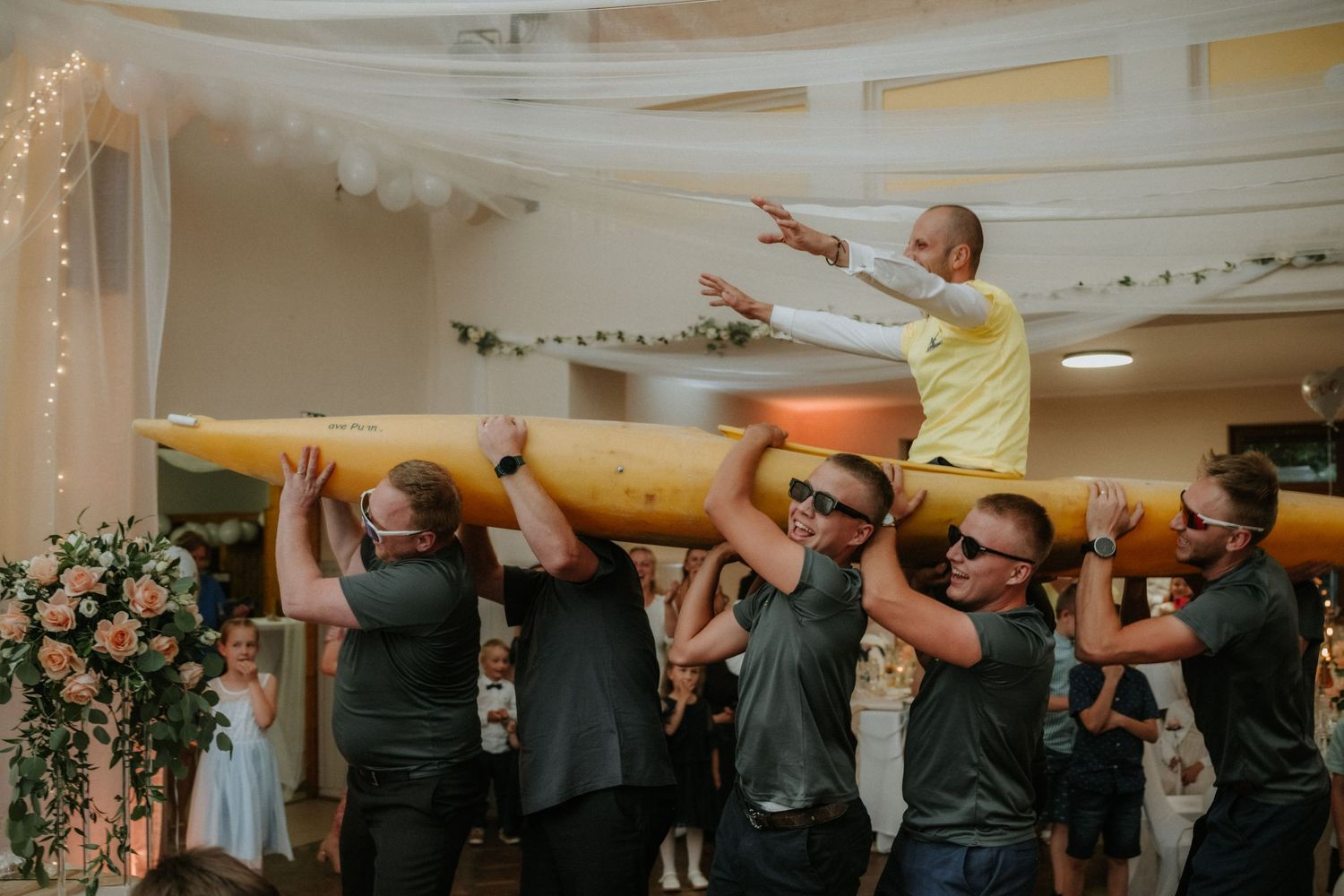 Men in dark shirts lift a person on a yellow log at a festive indoor celebration with floral decor.