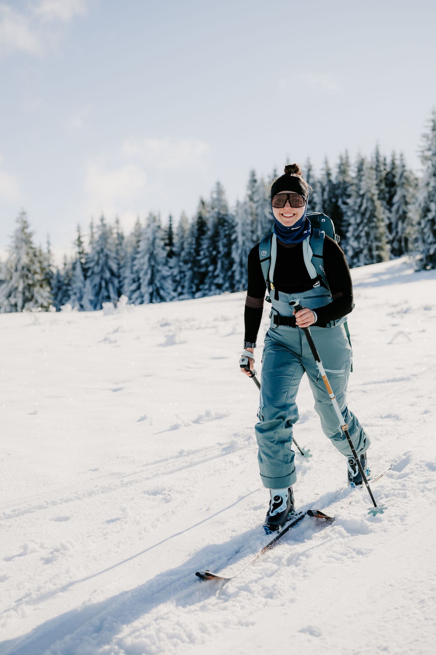 Smiling female skier in light blue pants and goggles ski touring near snow-covered pine forest.