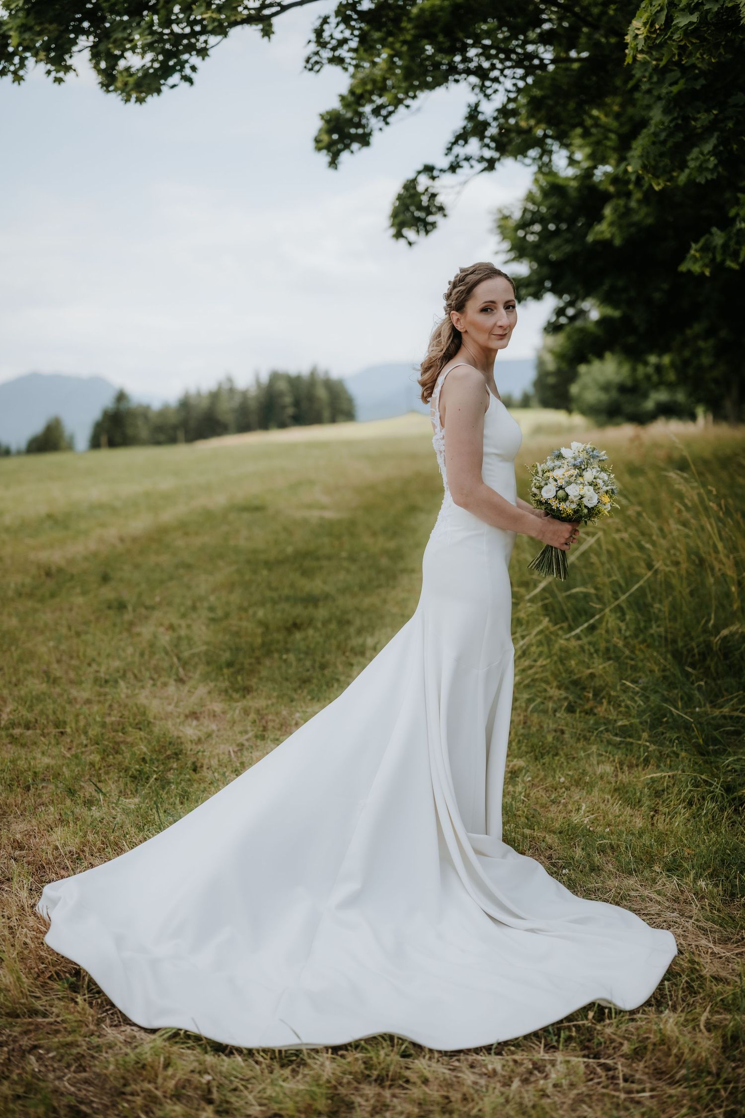 Bride in elegant white gown with train holds bouquet in scenic mountain meadow under oak tree.