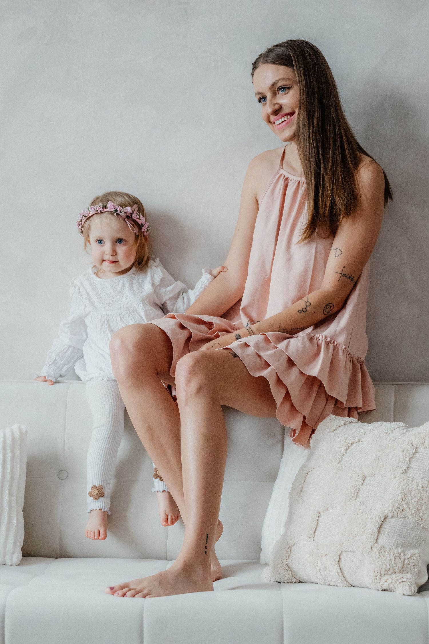 A mother in a pink dress sits on a white sofa beside her young daughter wearing a floral headband indoors.