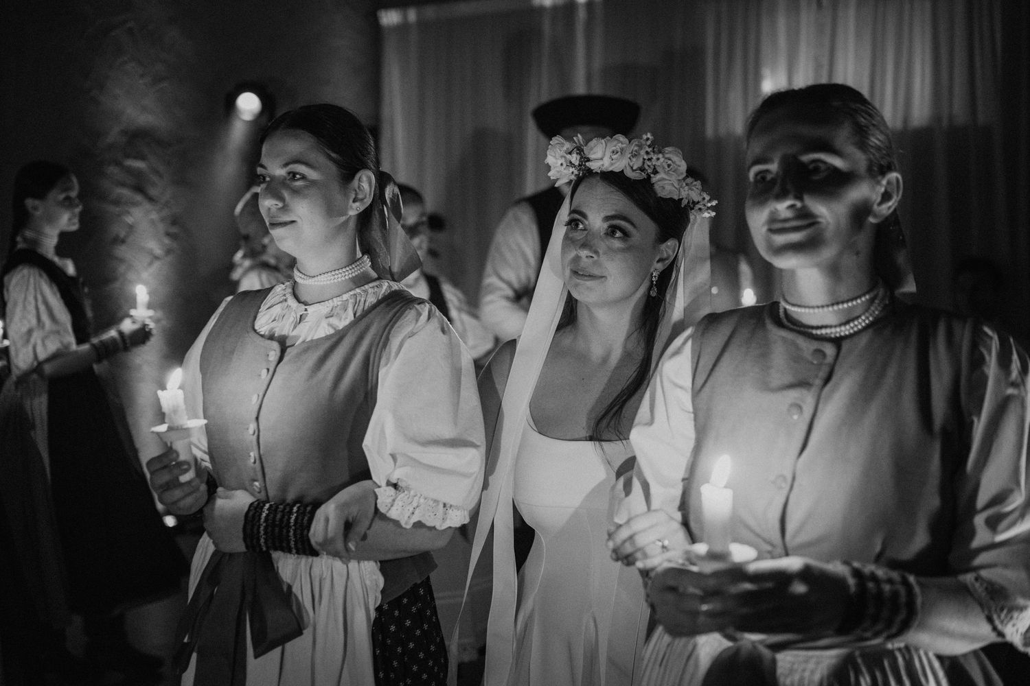 Black and white photo of women in traditional folk costumes holding candles during a ceremonial celebration.