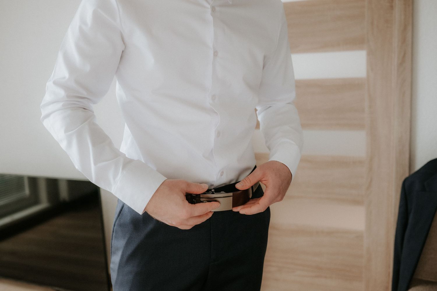 Man in white dress shirt fastening a black belt on dark trousers, standing near a wooden door.
