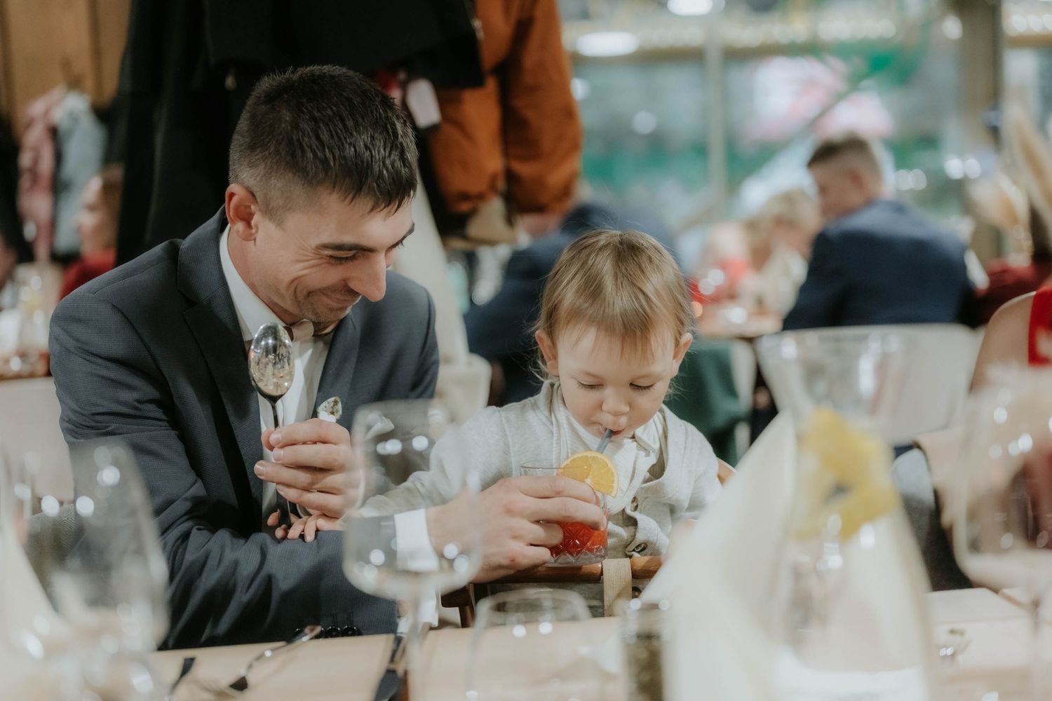 Man in gray suit sits with toddler at elegant wedding reception table with glassware.