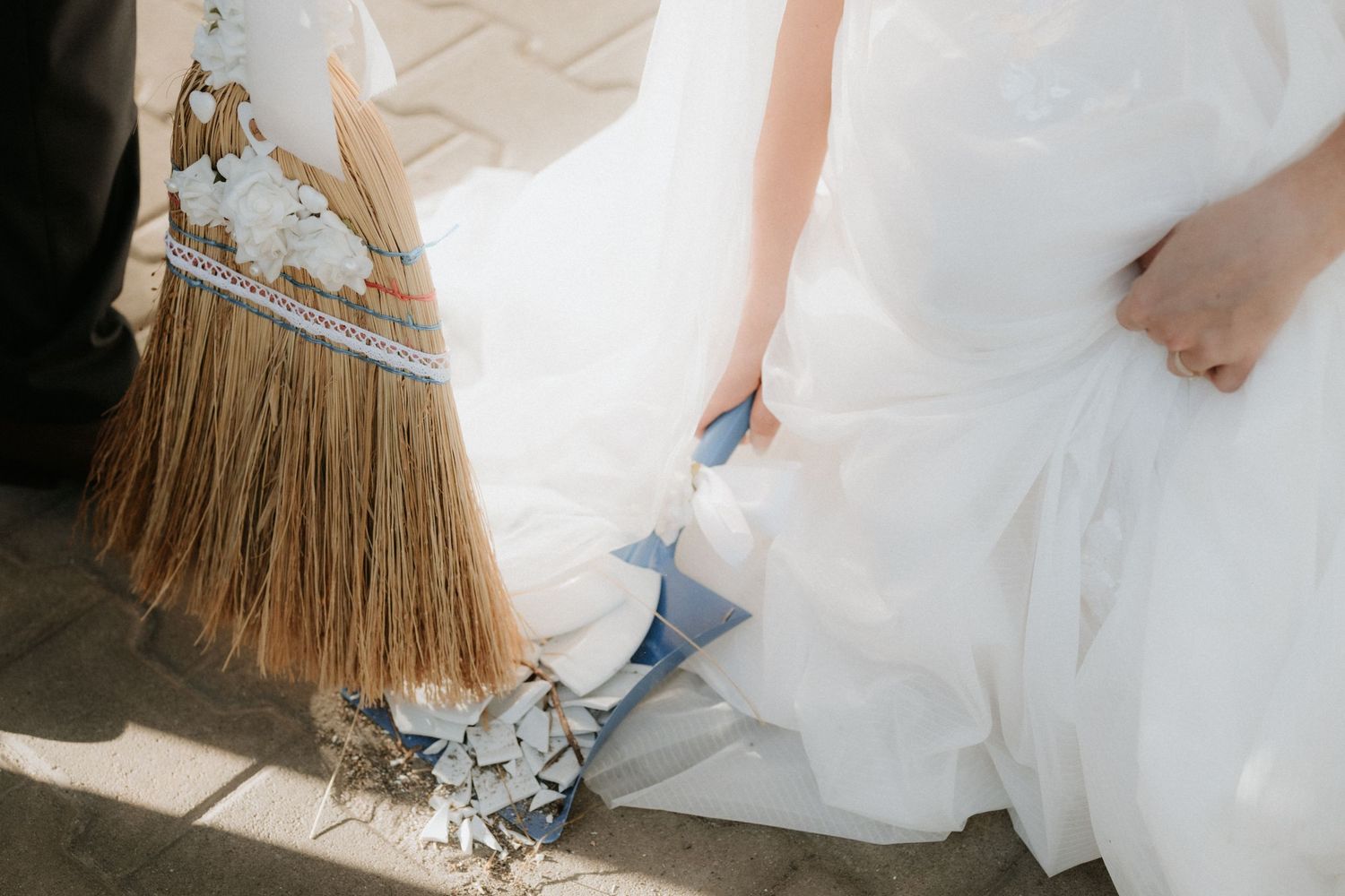Bride in white wedding dress holding floral bouquet next to decorative broom for jumping the broom ceremony.