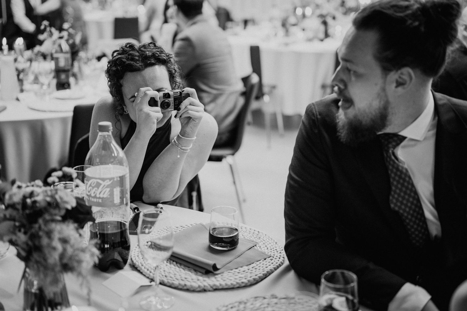 Black and white wedding reception photo of guests laughing, one taking a photo with a camera at a decorated table.