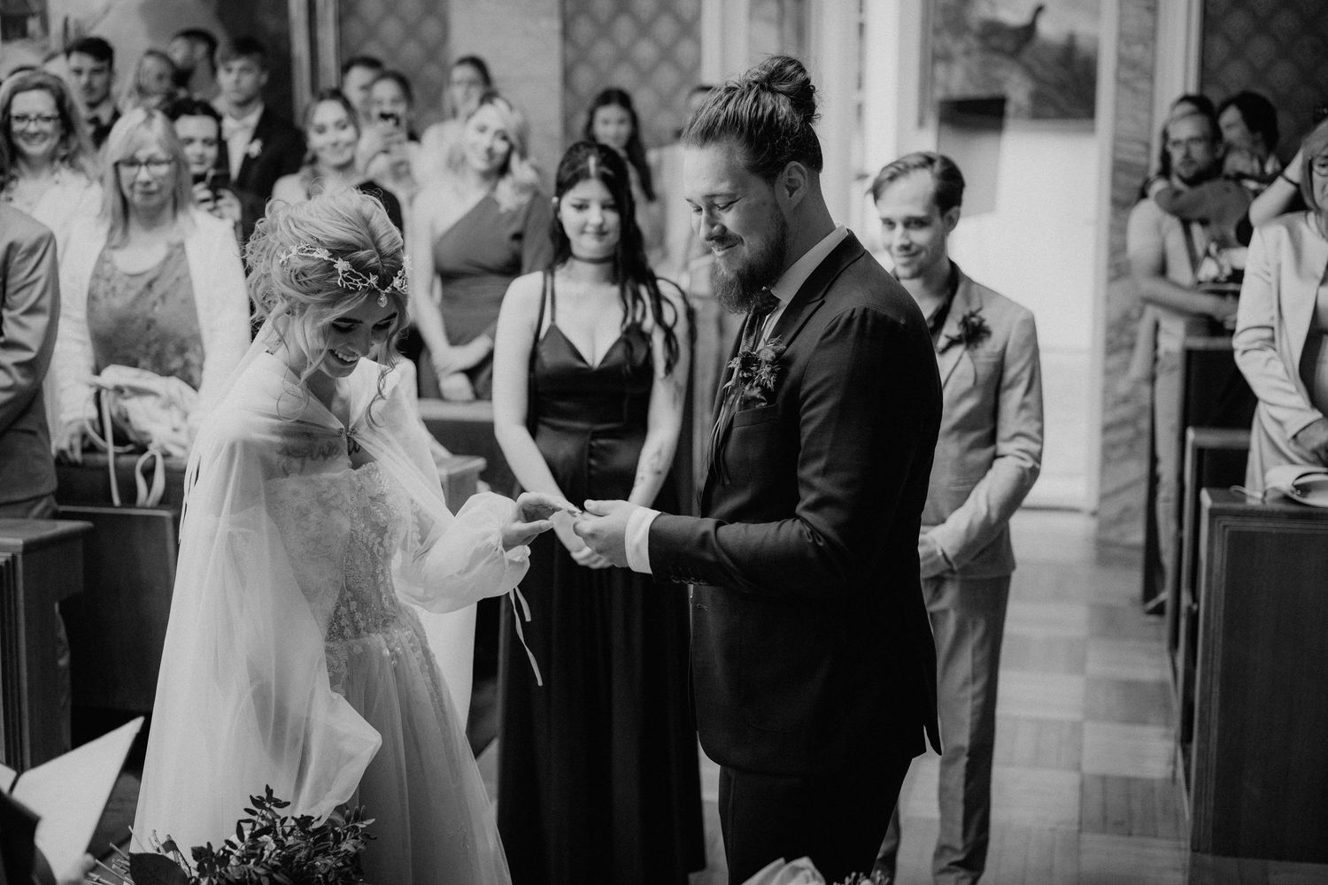 Black and white photo of a couple exchanging rings at their wedding ceremony with guests seated in pews behind them.