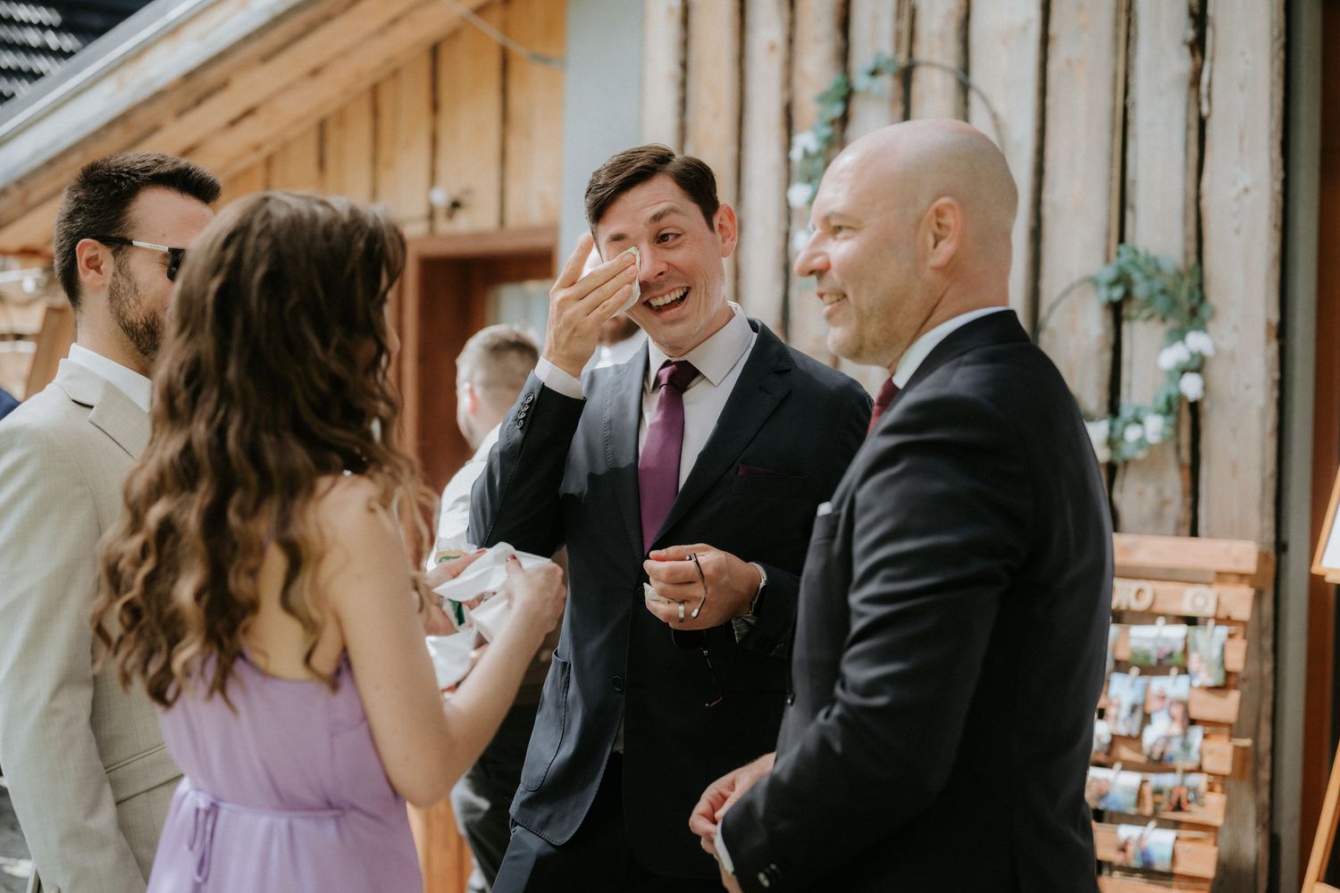 Guests laughing and socializing at a wedding reception, wearing formal attire in a rustic venue.