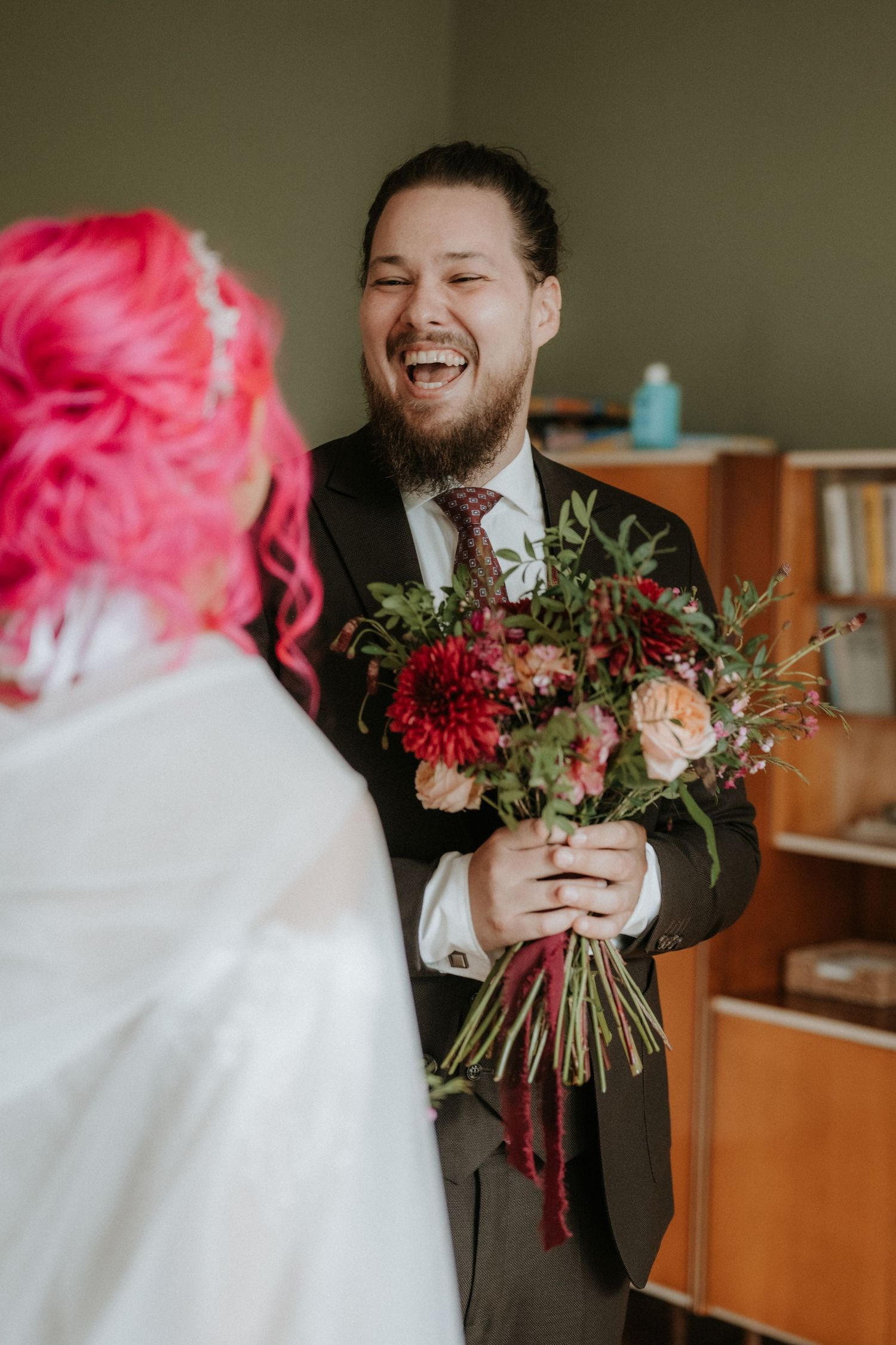 Groom laughing joyfully holding red and pink bridal bouquet during first look with pink-haired bride on wedding day.