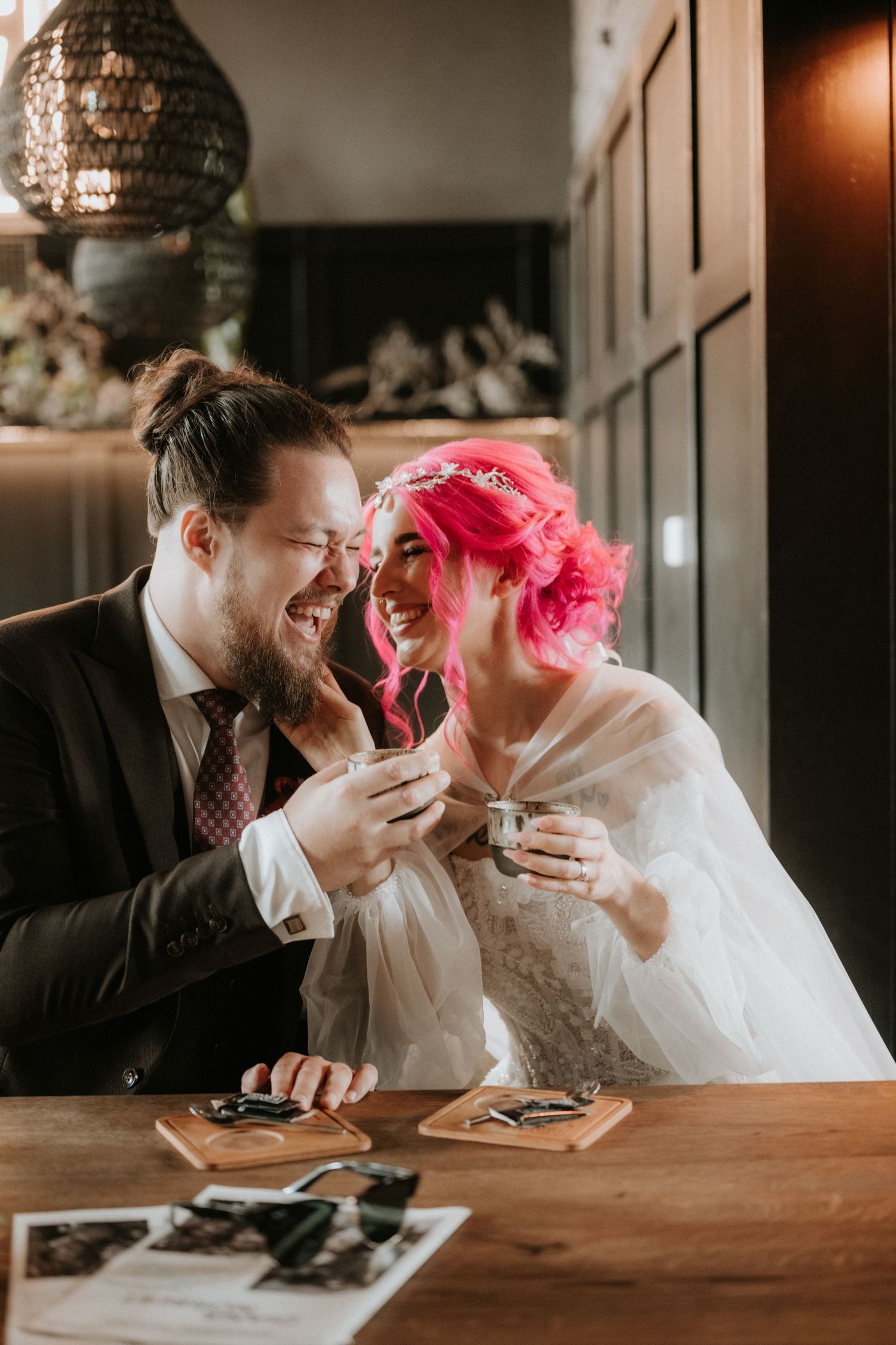 Bride with pink hair and groom in suit laughing together at a table, holding small cups during their wedding reception.