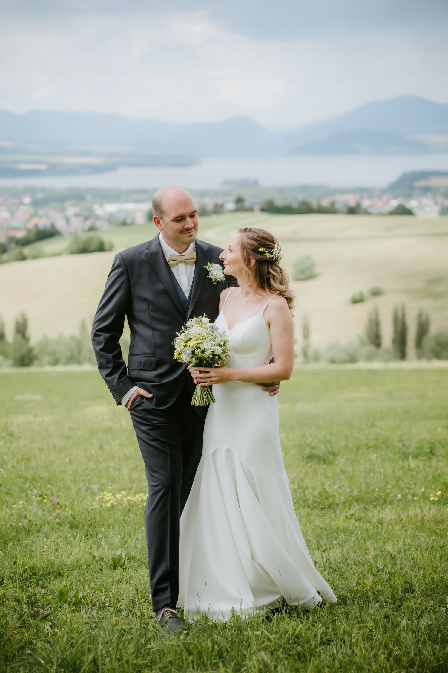 Bride and groom share a romantic moment on a green hillside with scenic lake and mountain views on their wedding day.