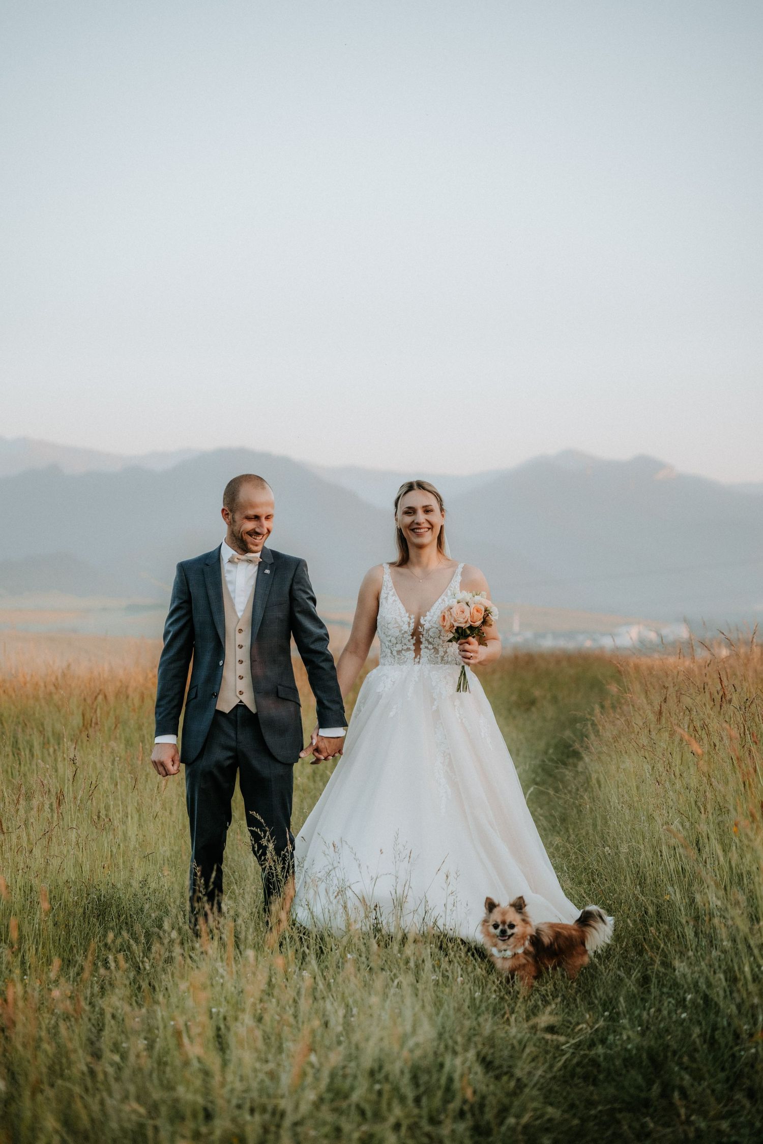 Bride and groom holding hands in a meadow with mountains, bride holding bouquet, small dog beside them.