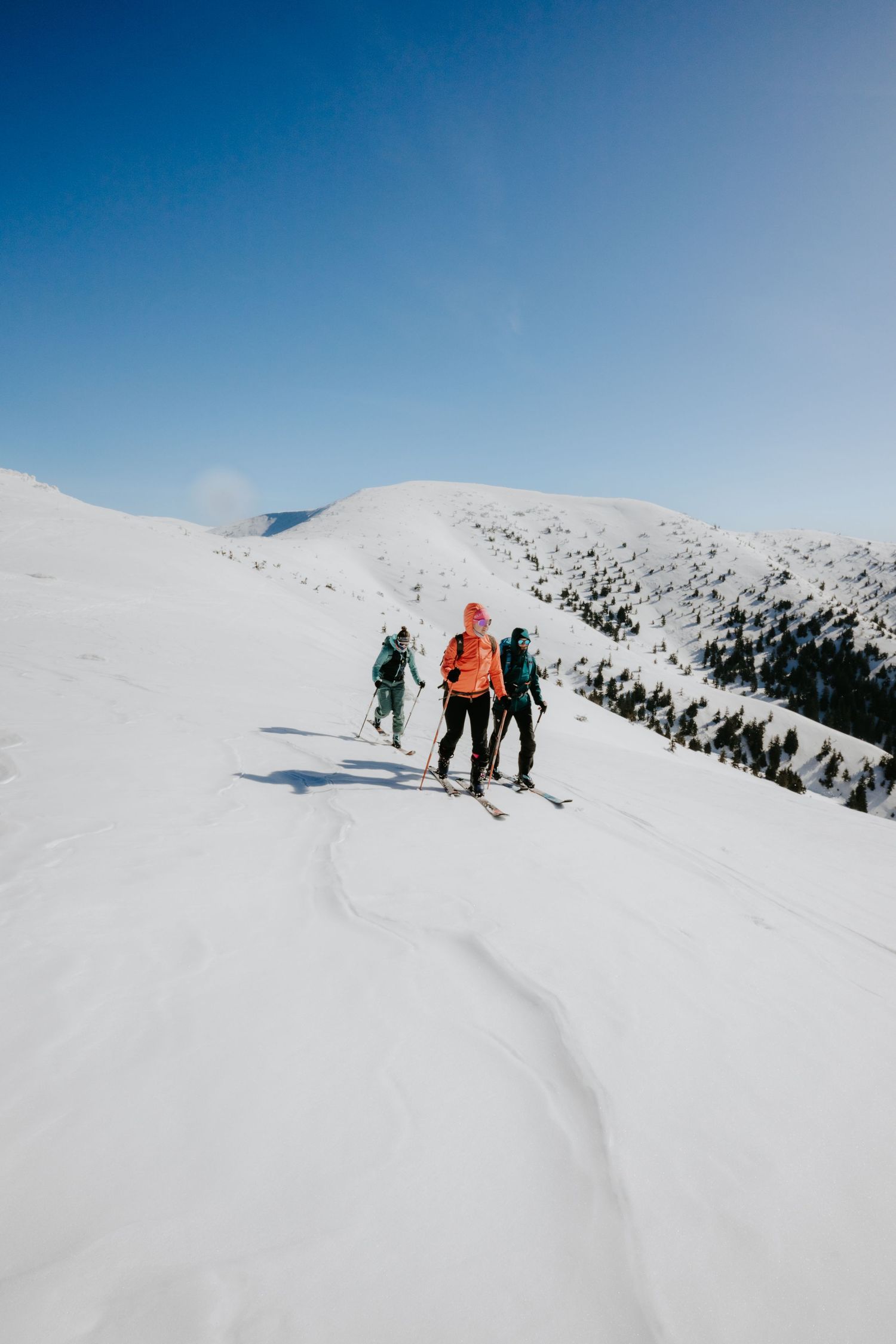 Three backcountry skiers with colorful backpacks ascending a snow-covered mountain ridge on a clear day.