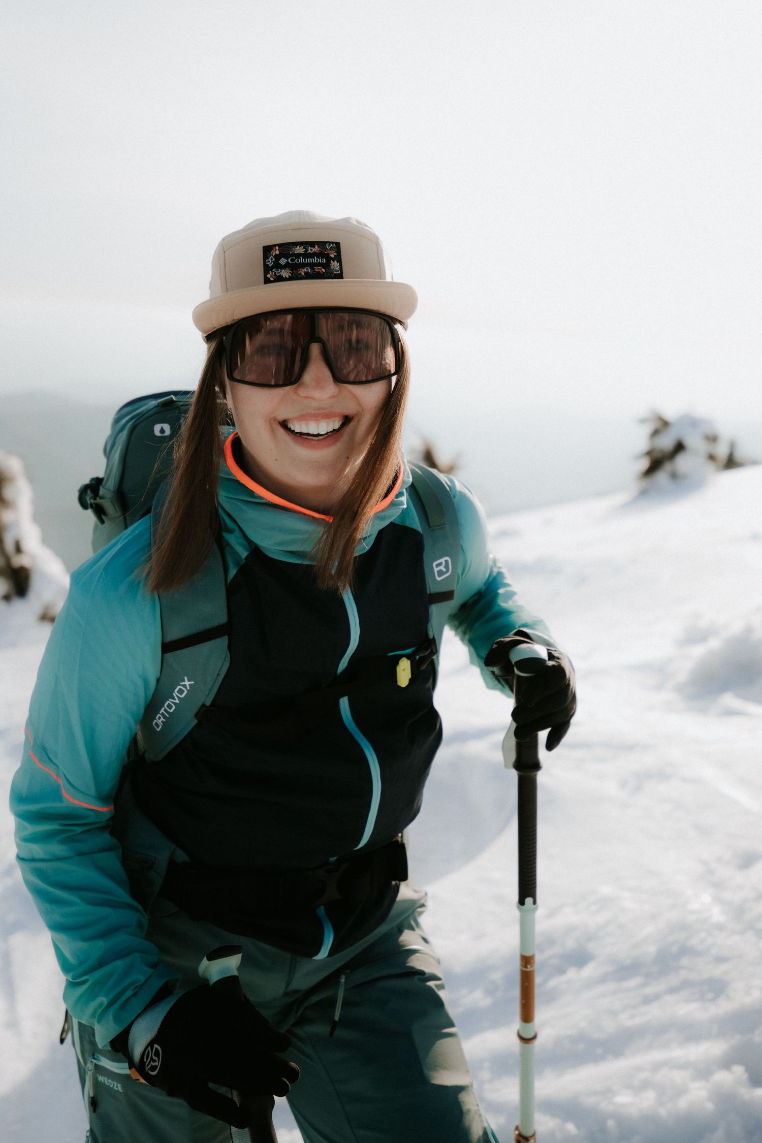 Smiling female skier in teal Ortovox jacket and tan cap posing with ski pole on a snowy mountain.