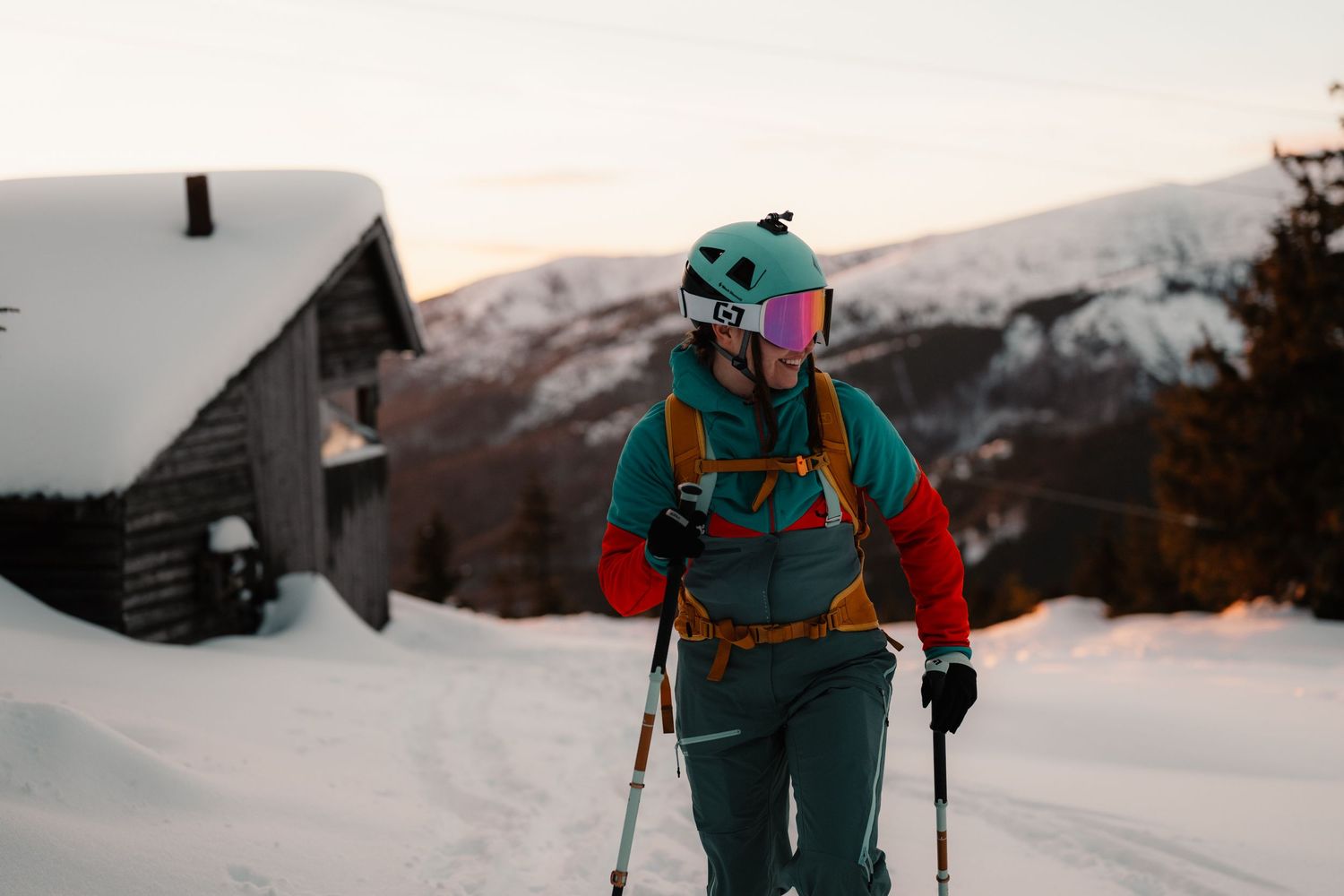 Female backcountry skier with poles walking through deep snow past rustic cabin at sunset.