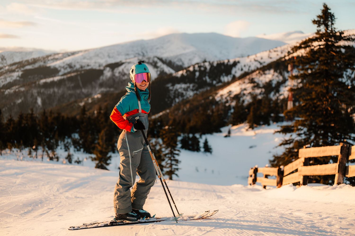 Female skier standing on snowy mountain slope at golden sunset with ski poles and colorful gear.
