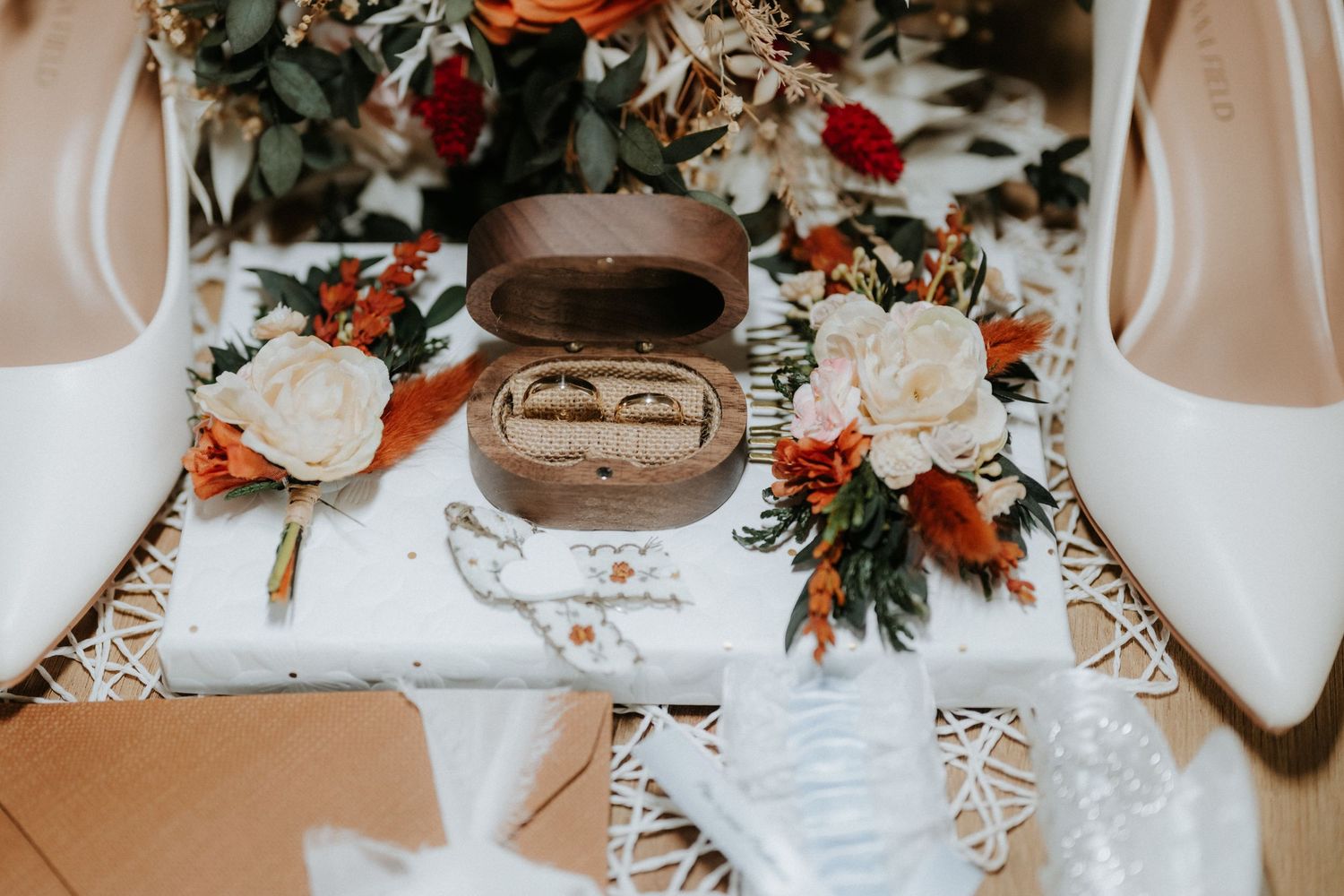 Wedding rings in wooden box surrounded by white shoes, floral bouquet, and bridal accessories.