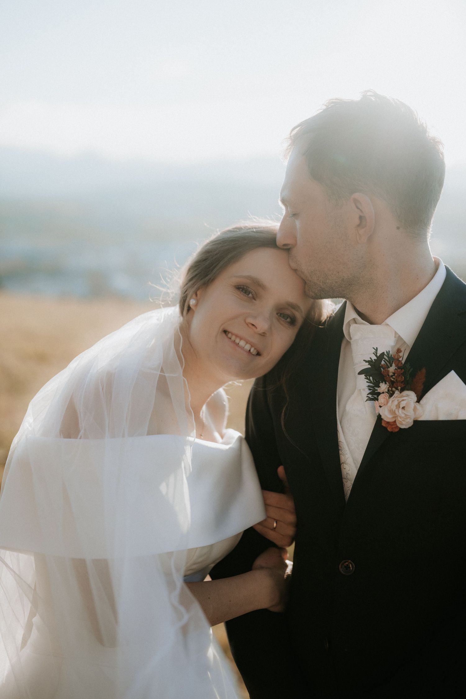 Groom kissing bride on the forehead while she smiles on their sunny outdoor wedding day.