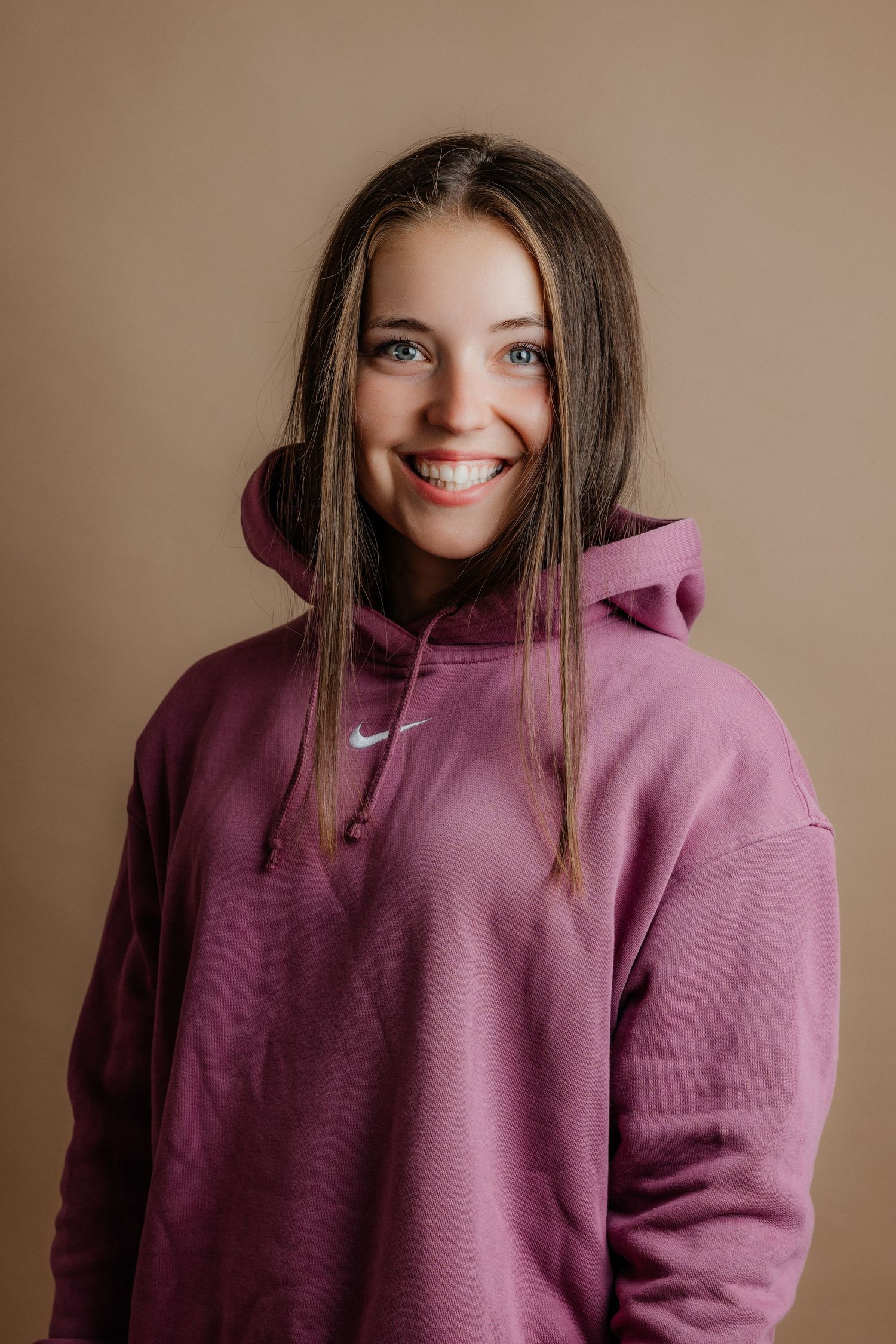 Happy young woman wearing oversized pink Nike hoodie smiling against warm beige studio background.