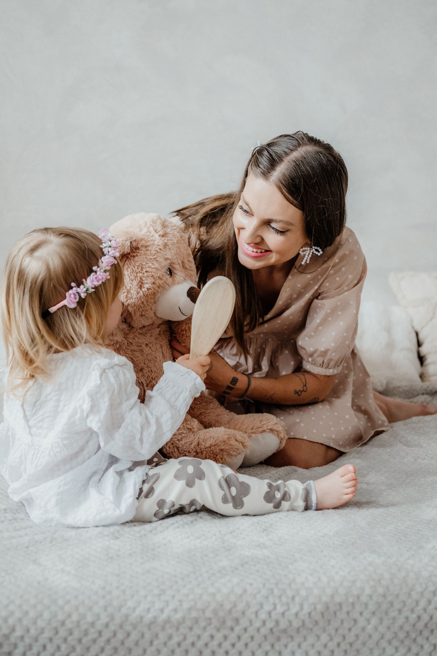Mother and young daughter brushing a large teddy bear together on a cozy bed.