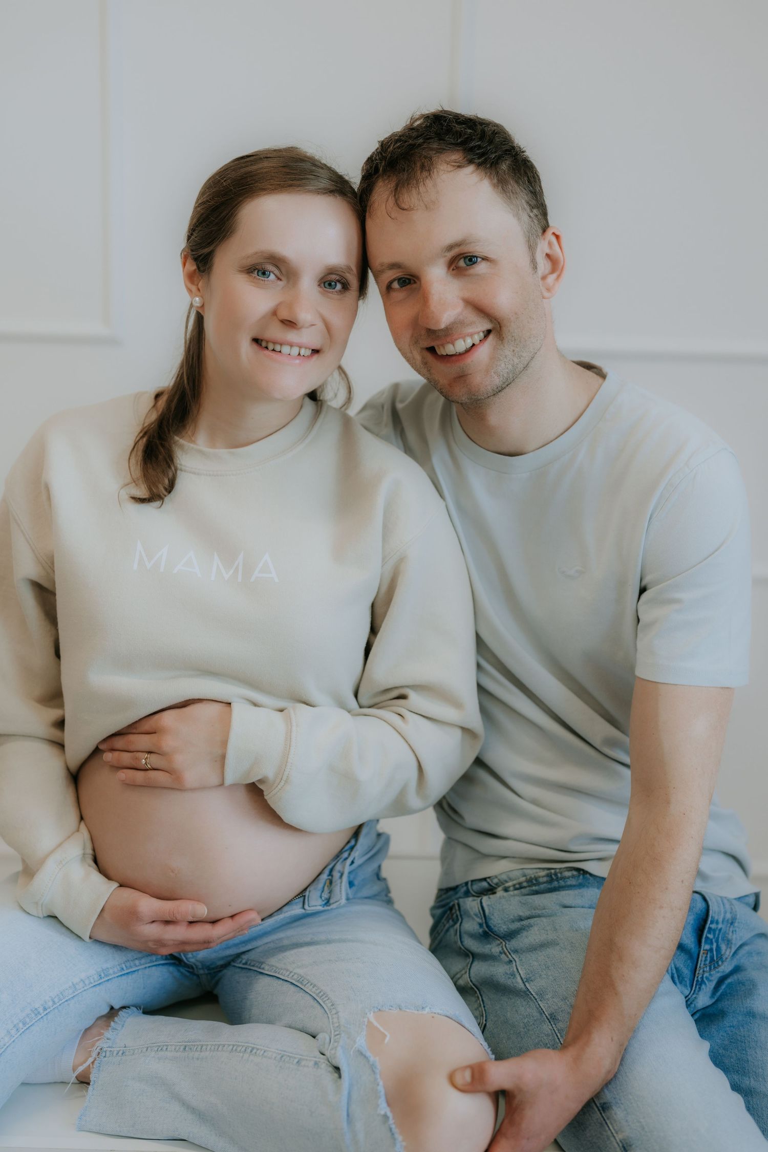 Happy expecting couple in matching beige MAMA sweatshirts smiling at camera during maternity session.