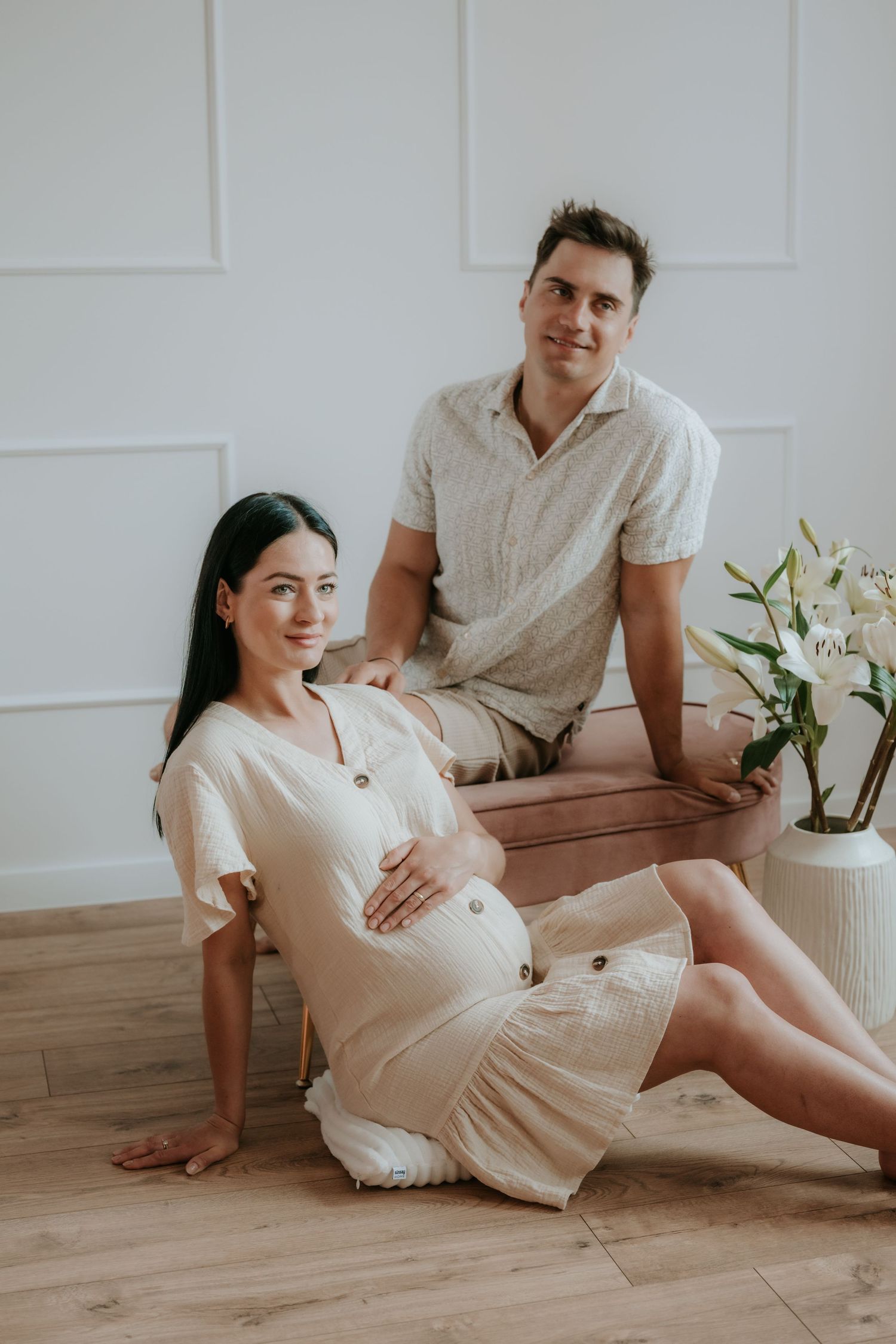 Pregnant couple posing in bright studio, woman in cream dress sitting on floor, man behind her.