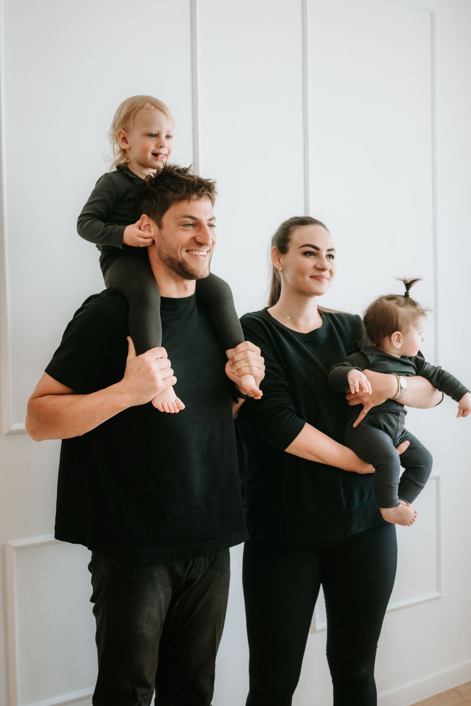 Smiling family in black clothing, father with toddler on shoulders and mother holding baby indoors.