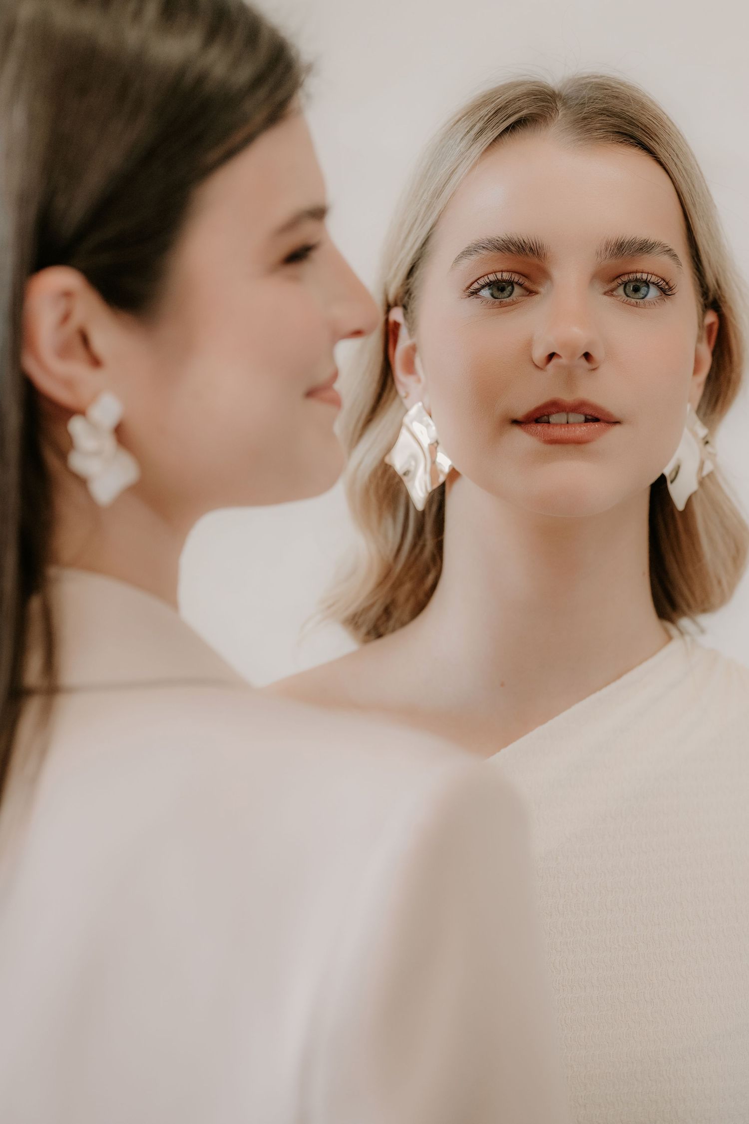 Two women wearing statement white earrings, one blonde and one brunette, in soft-focus portrait.