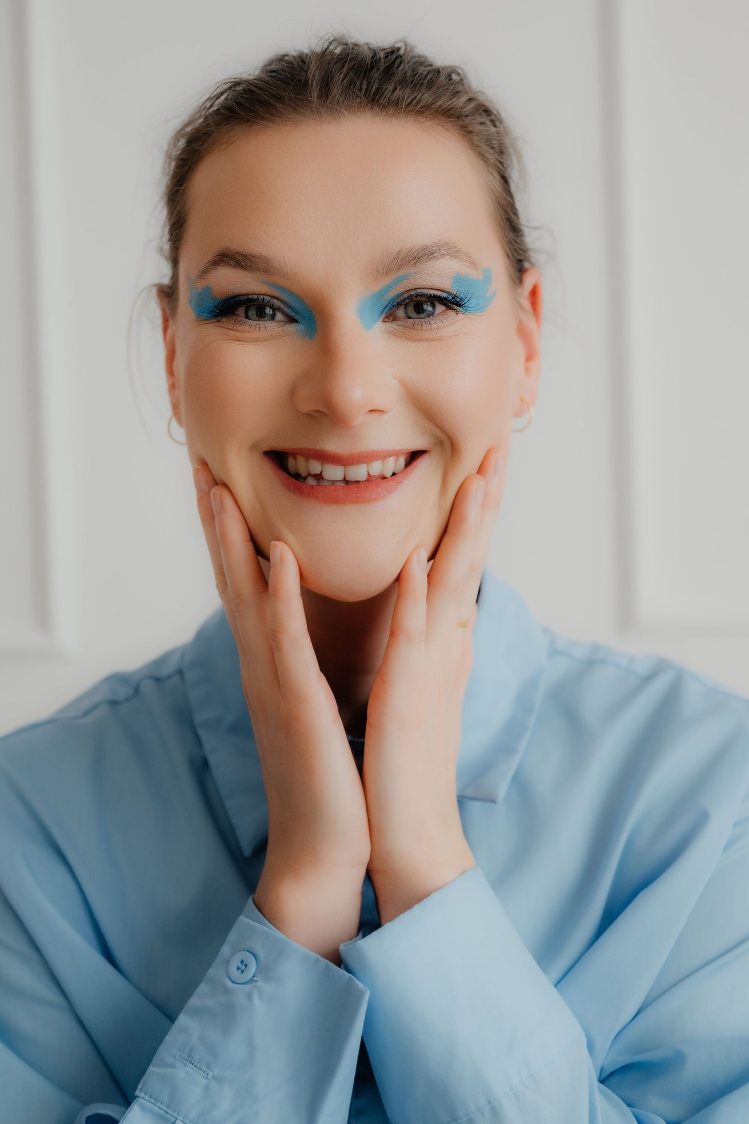 Happy woman with bold blue eyeshadow in blue shirt smiling with hands on cheeks.