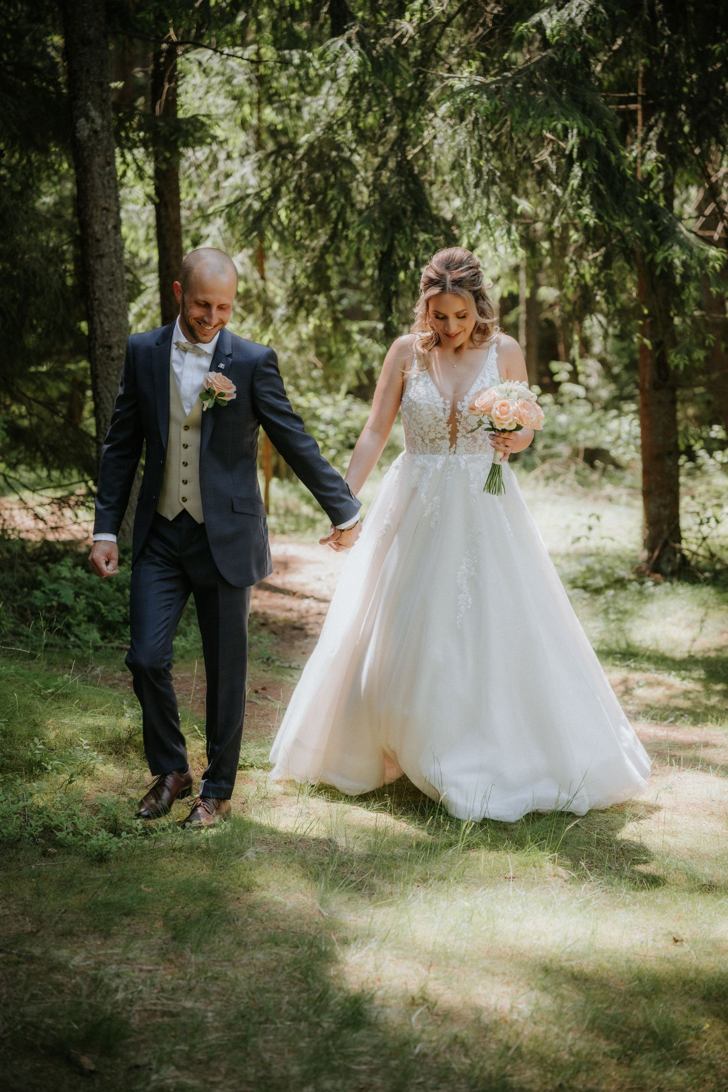 A bride and groom walk hand in hand through a sunlit forest, she in a white gown holding a bouquet, he in a navy suit.