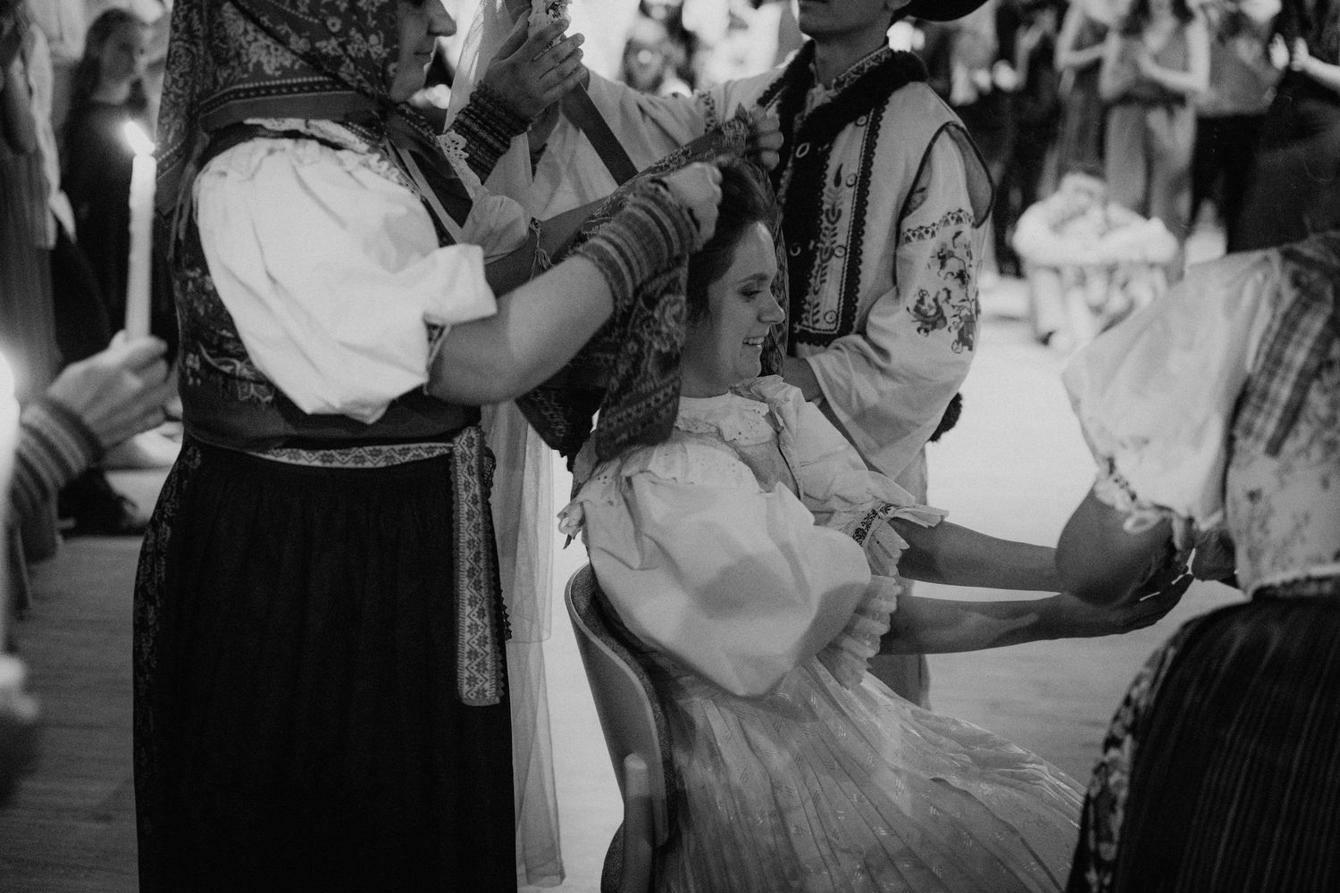 Women in traditional folk costumes perform a ceremonial hair braiding ritual indoors.