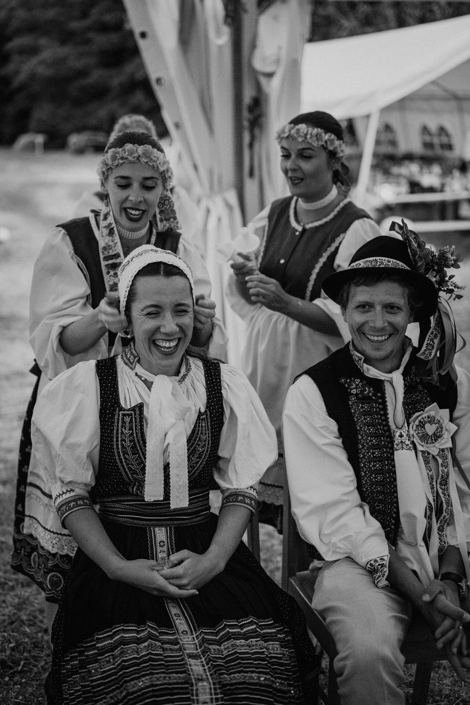 Black and white photo of people in traditional folk costumes laughing joyfully at an outdoor cultural celebration.