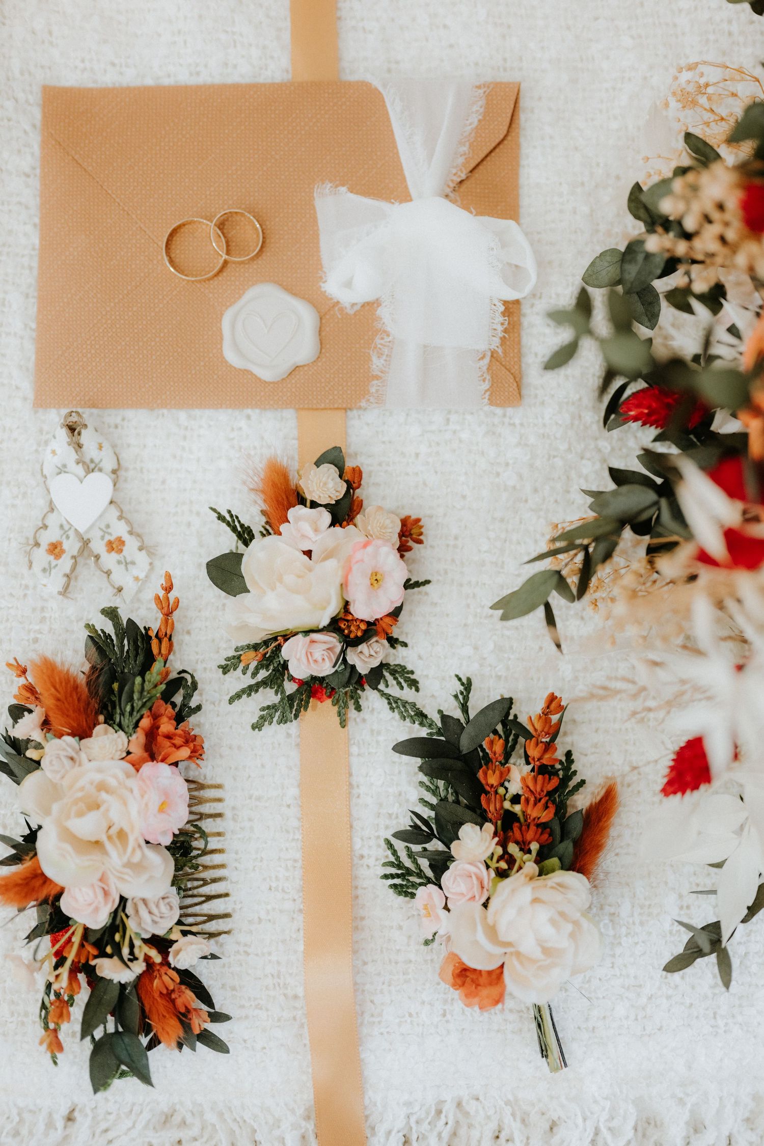 Elegant wedding flat lay with gold rings, floral boutonnieres, hair comb, kraft envelopes, and lush blooms in warm terracotta tones.