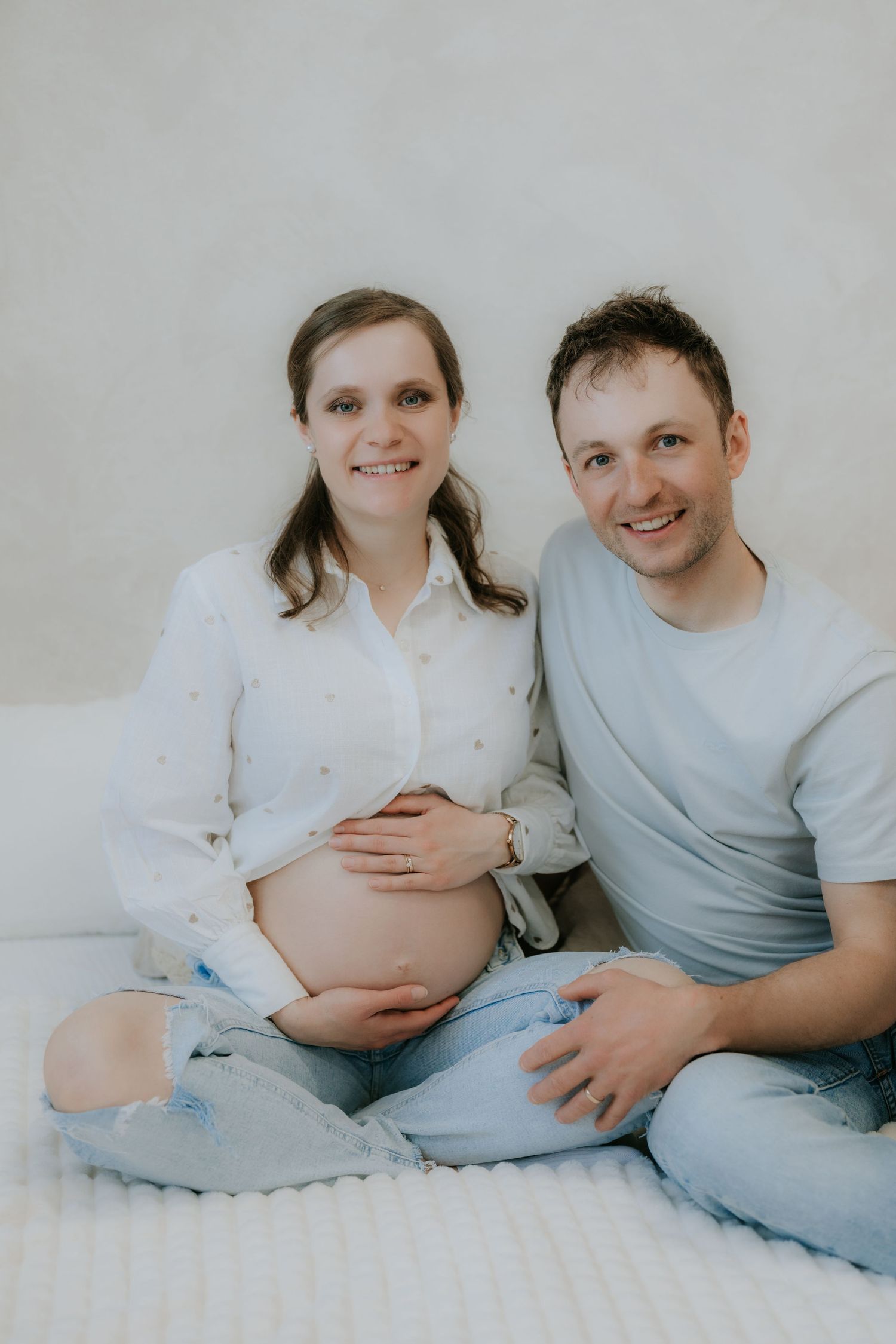 Happy expectant couple sitting together on white surface, smiling at camera during maternity shoot.