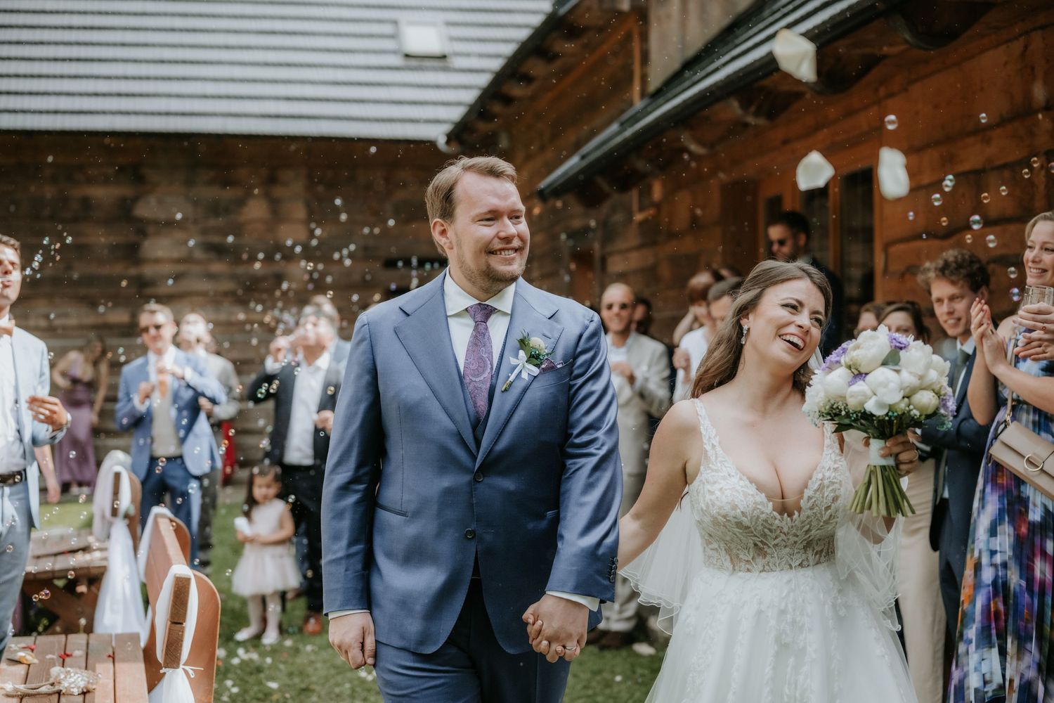 Happy newlyweds smile while walking through bubble send-off at rustic outdoor wedding ceremony.