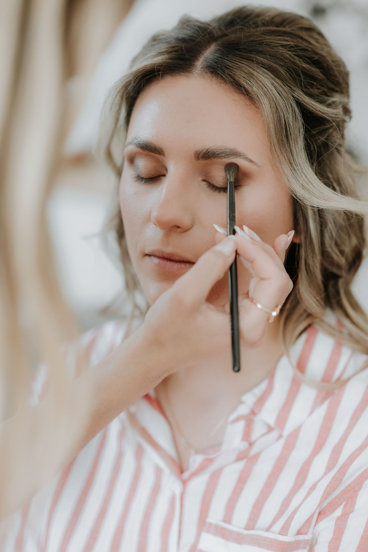 Makeup artist applying eyeshadow to a bride in a pink striped robe during wedding preparations.