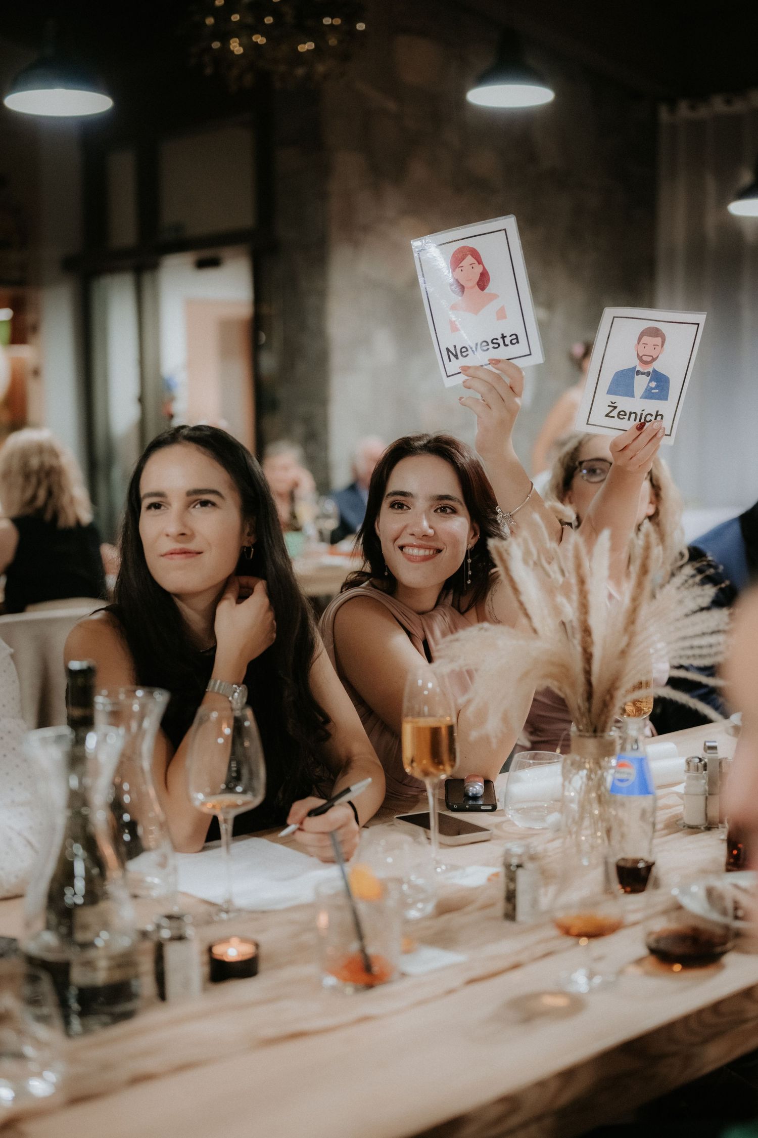Women holding bride and groom cards at a wedding reception table with drinks and floral decor.