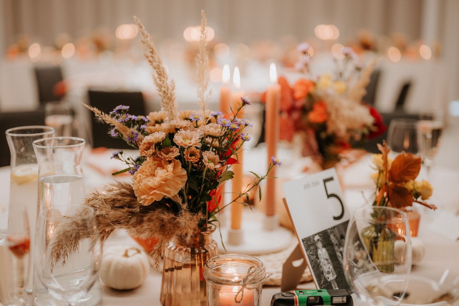 Elegant fall wedding reception table with pampas grass, orange floral arrangements, candles, and a table number 5 sign.