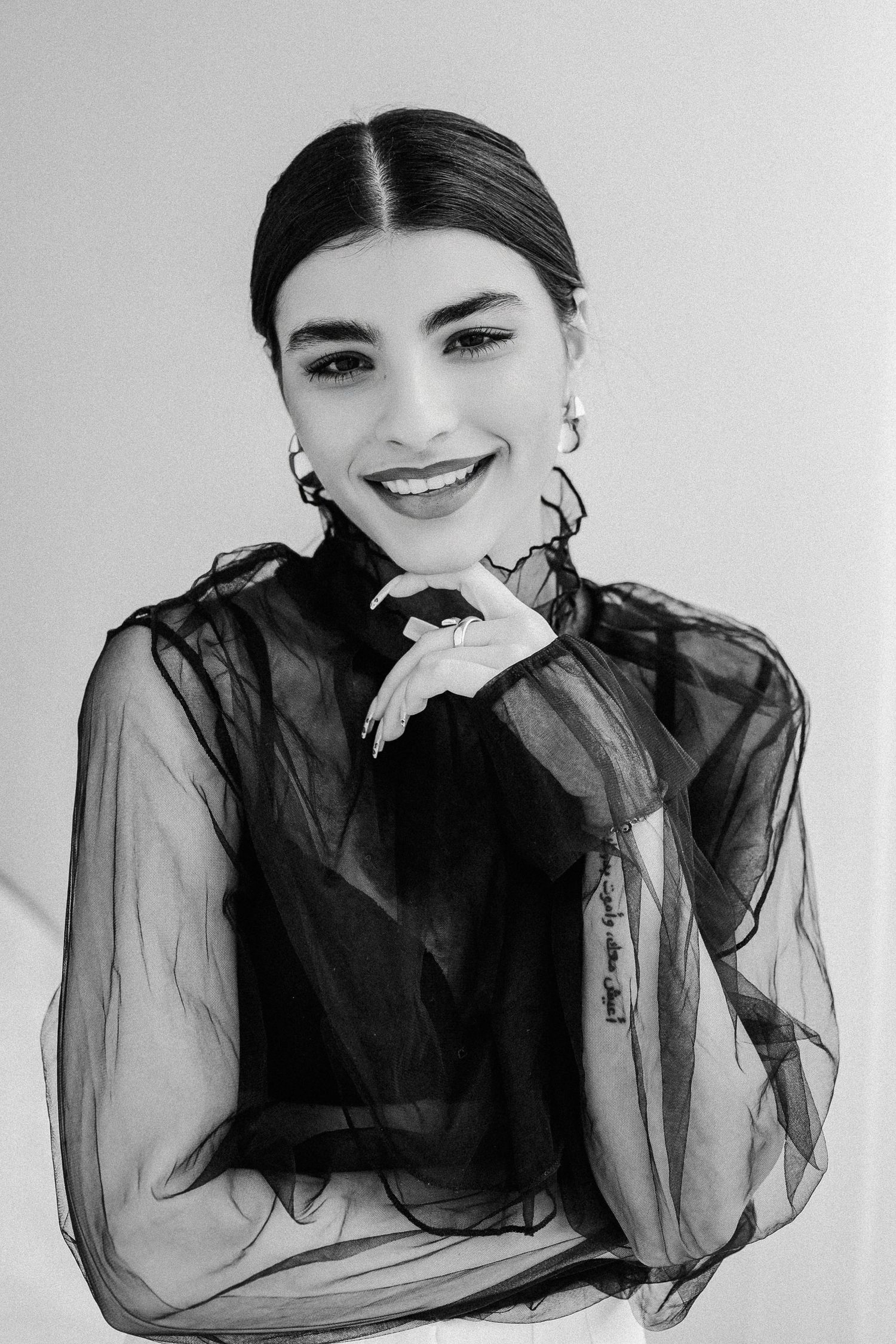 Black and white portrait of smiling woman in sheer black blouse with hoop earrings.