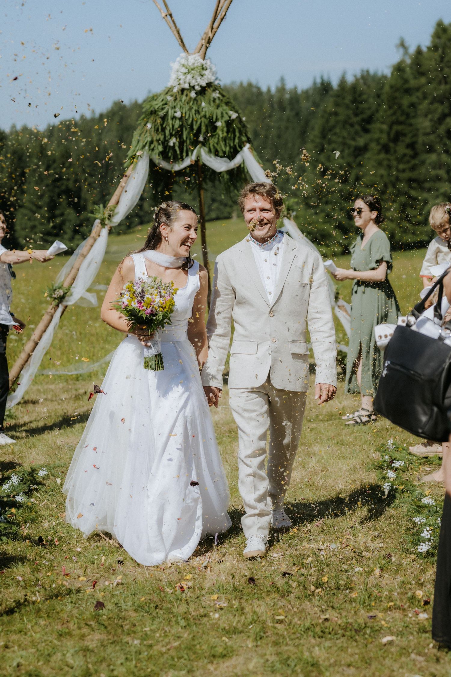 Joyful newlyweds walk through confetti shower at outdoor wedding ceremony with floral arch and forest backdrop.