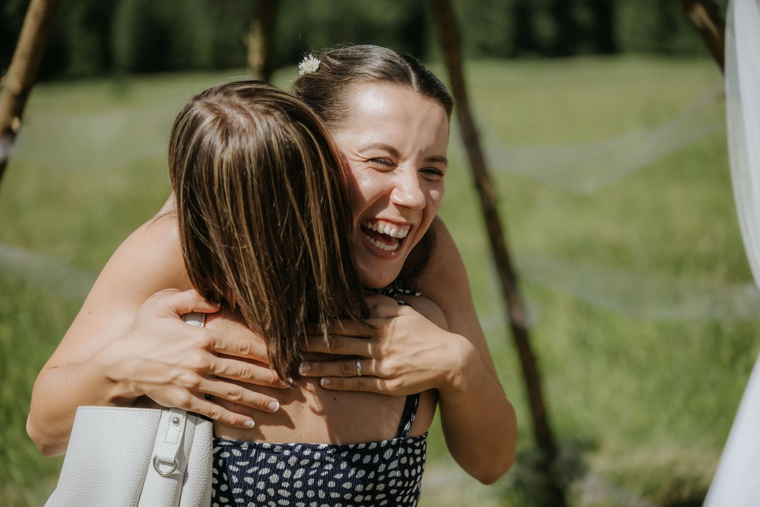 Two women sharing a joyful hug outdoors at a wedding celebration in a green meadow setting.