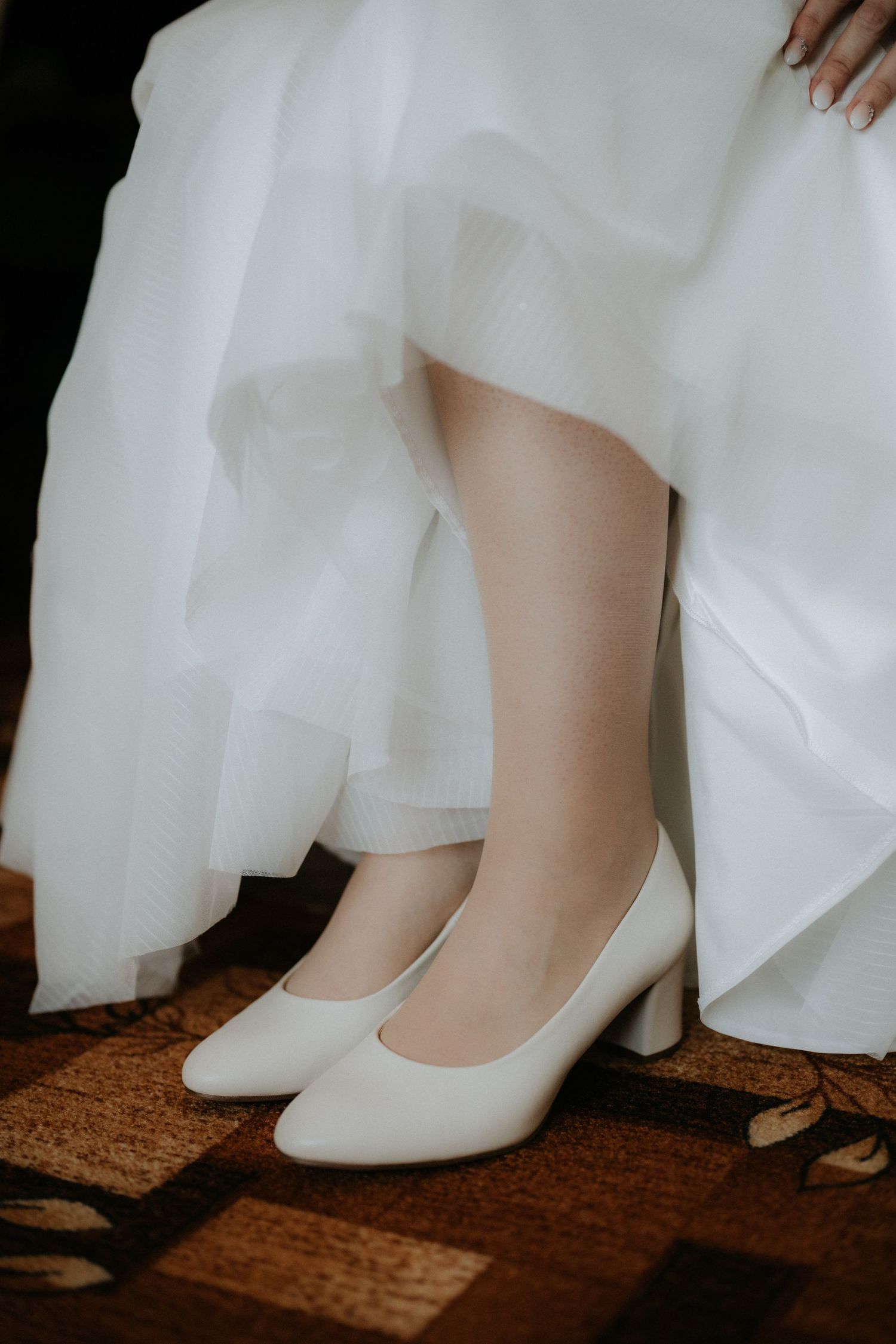 Bride wearing elegant white block heel pumps peeking beneath a flowing tulle wedding dress while seated.