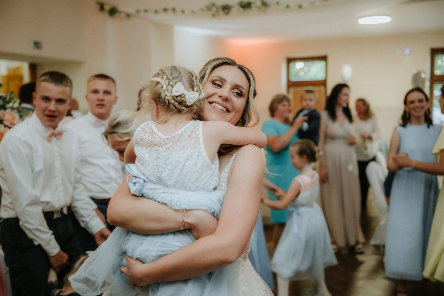 Bride in white dress hugs a young flower girl at a wedding reception with guests in the background.