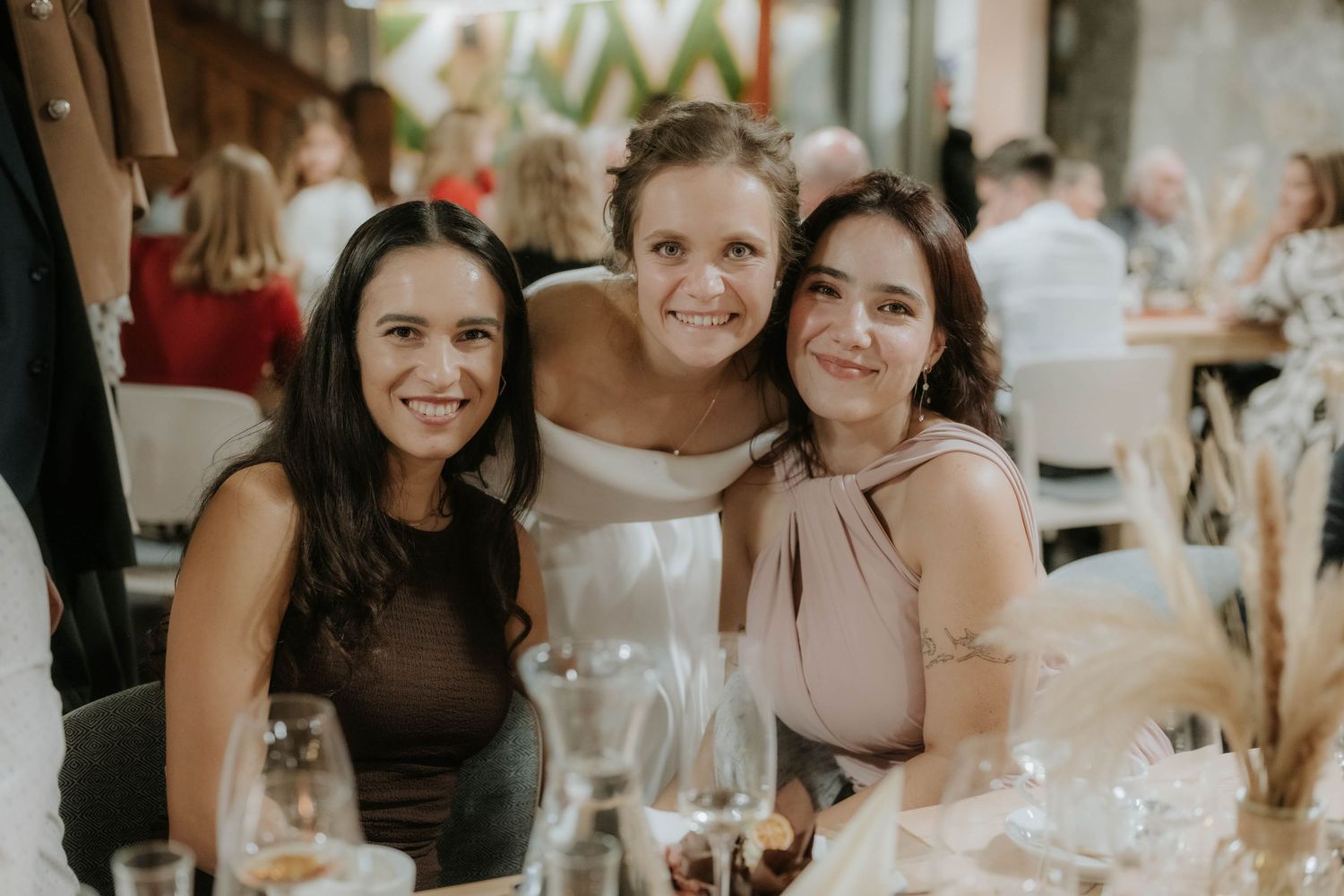 Three women smiling together at a wedding reception, with the bride in a white dress in the center.