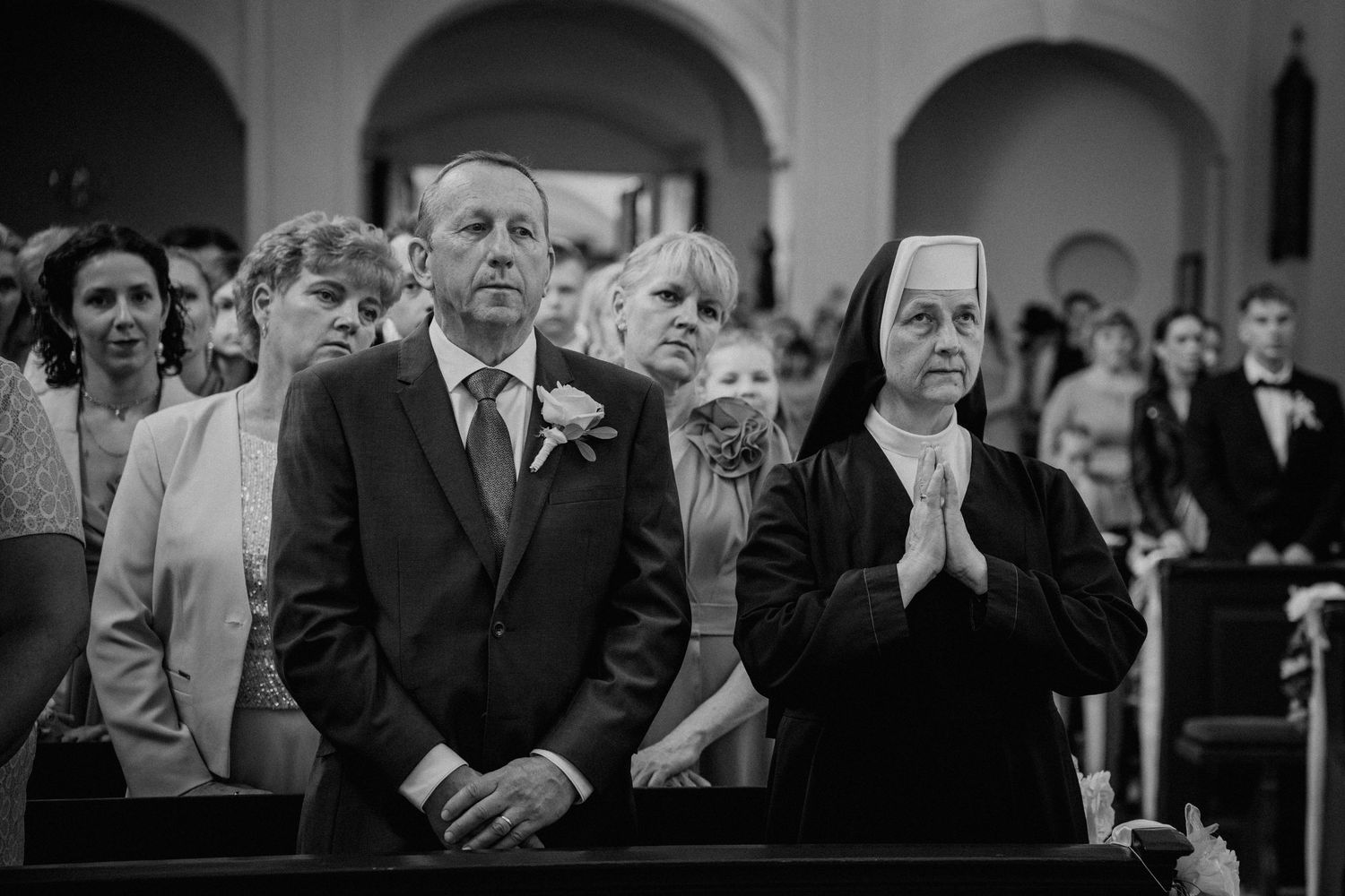 Black and white photo of a man in suit and nun praying in church pew during a wedding ceremony.