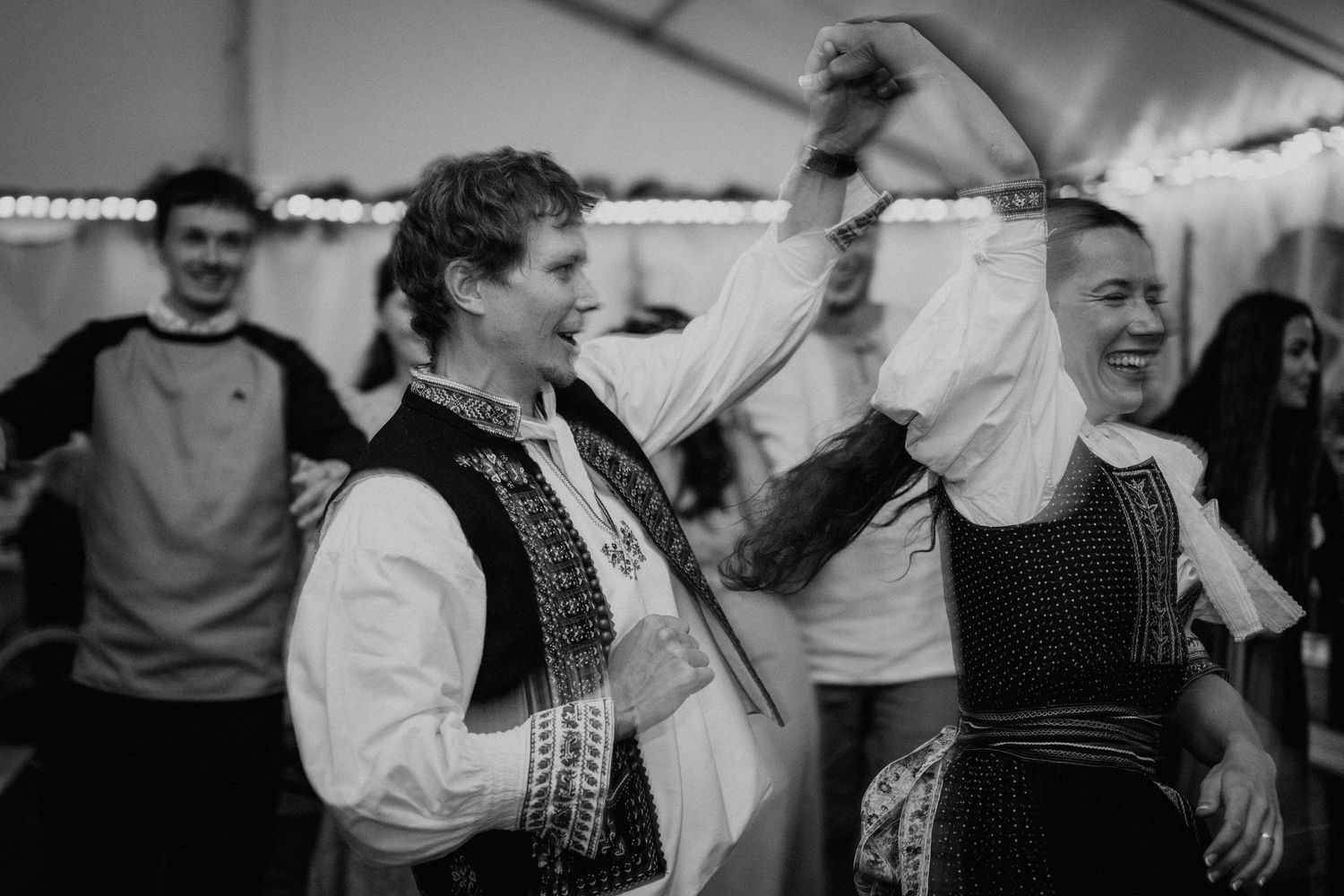 Black and white photo of people in traditional folk costumes dancing joyfully at a cultural celebration.