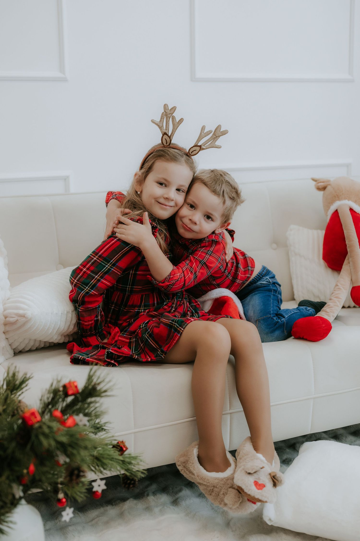 Two young children in red plaid outfits with reindeer antler headbands hugging on white sofa at Christmas.