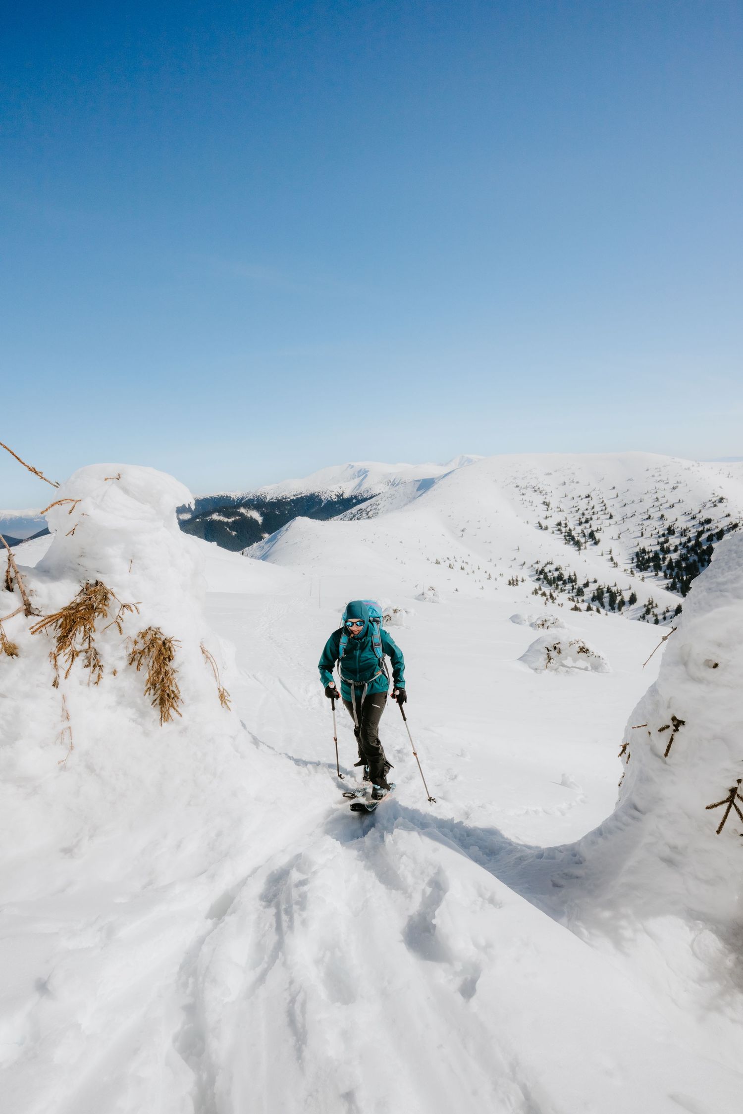 Female skier in teal jacket climbing a steep snowy mountain trail with ski poles on a clear day.