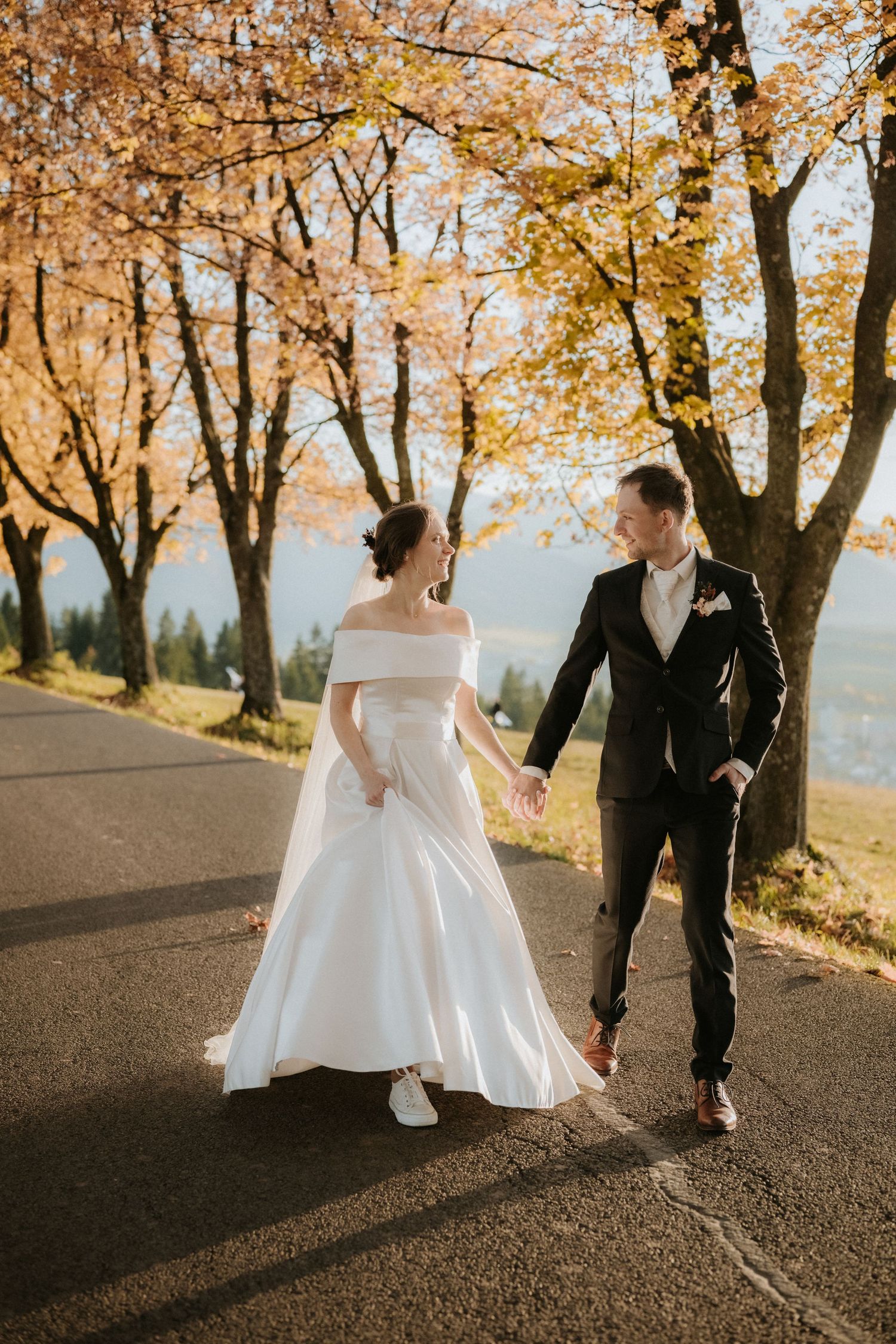 Bride and groom walk hand-in-hand on a sunlit road surrounded by golden autumn trees in a romantic wedding photo.