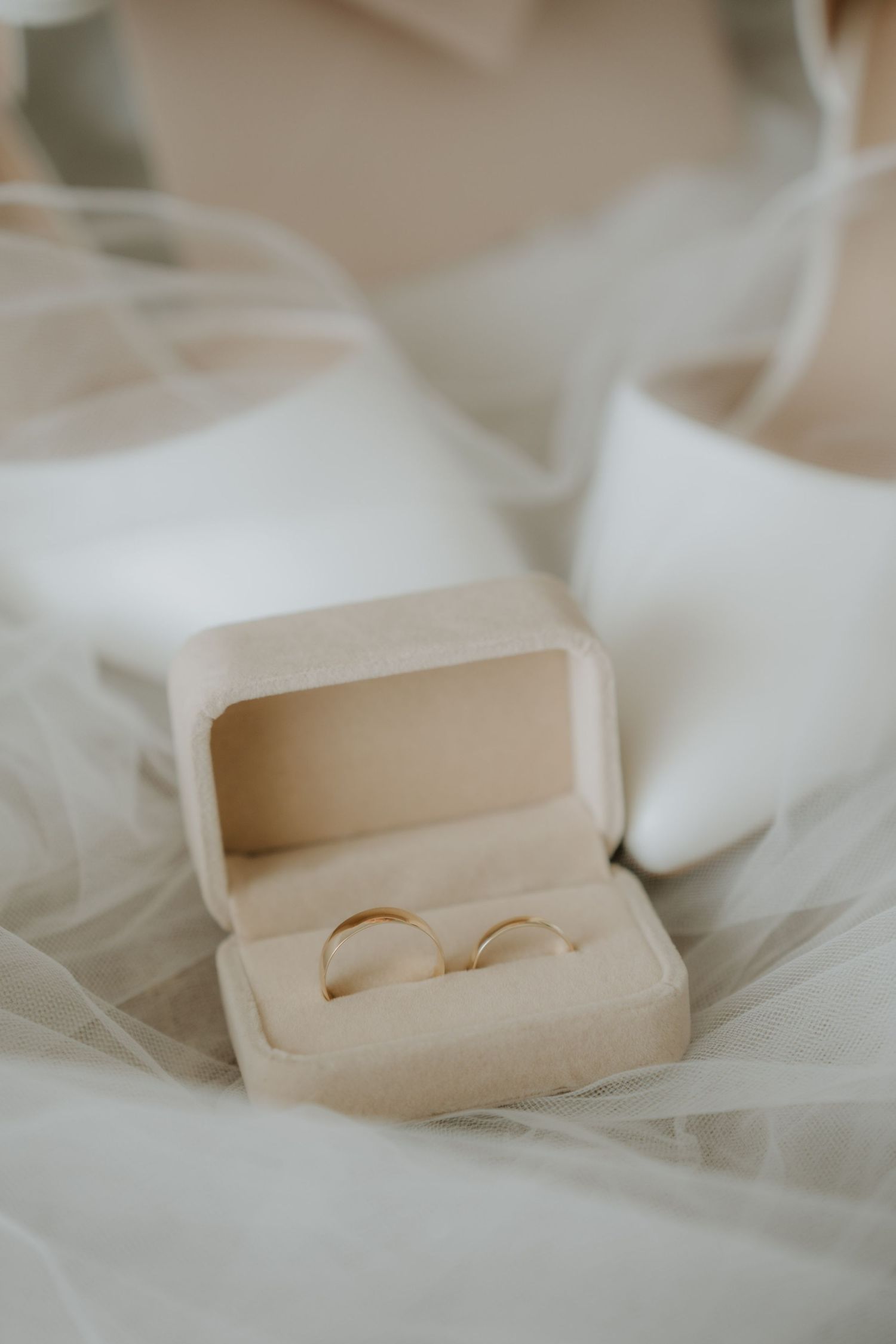 Two gold wedding bands displayed in a beige velvet ring box on a white bridal veil with satin heels.